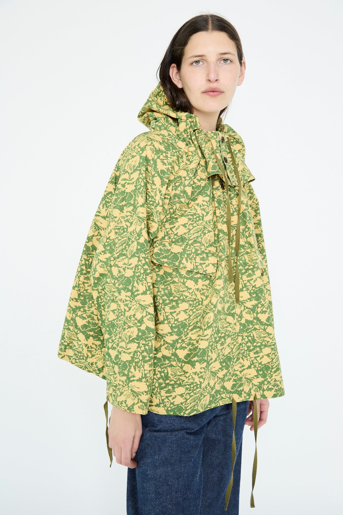 Dressed in Chimala's Summer Leaf Smock Jacket in Camo, a model pairs the oversized green and yellow hooded outerwear with jeans against a white background.