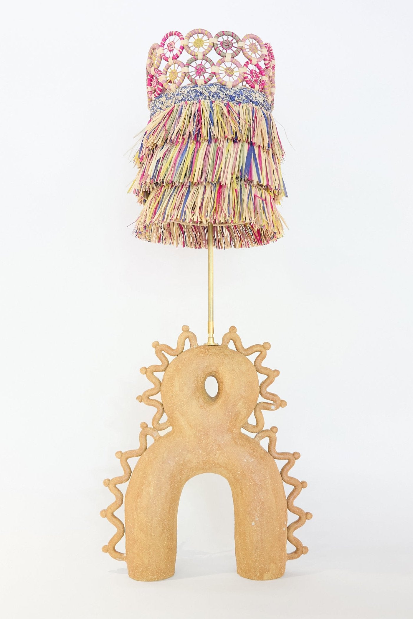 The Mora Mora Lamp with Multicolor Raffia Shade by Clandestine showcases a tan base with arch-like shapes, adorned with a tiered, colorful raffia lampshade and fringed structure.