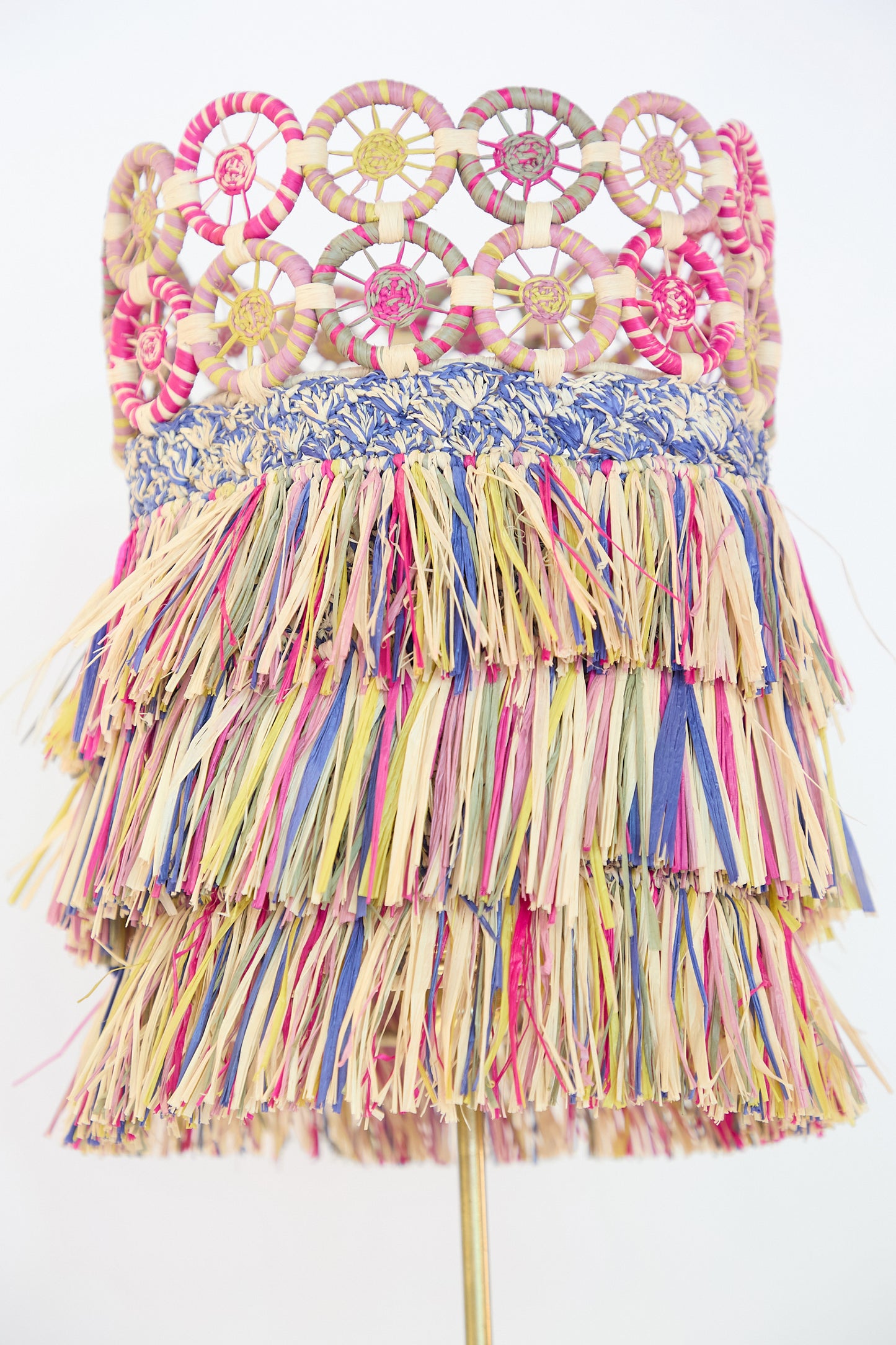 A hat featuring a top of circular woven patterns and layered multicolor raffia tassels below, reminiscent of Clandestine's Mora Mora Lamp with Multicolor Raffia Shade with its artful touches, on a white background.