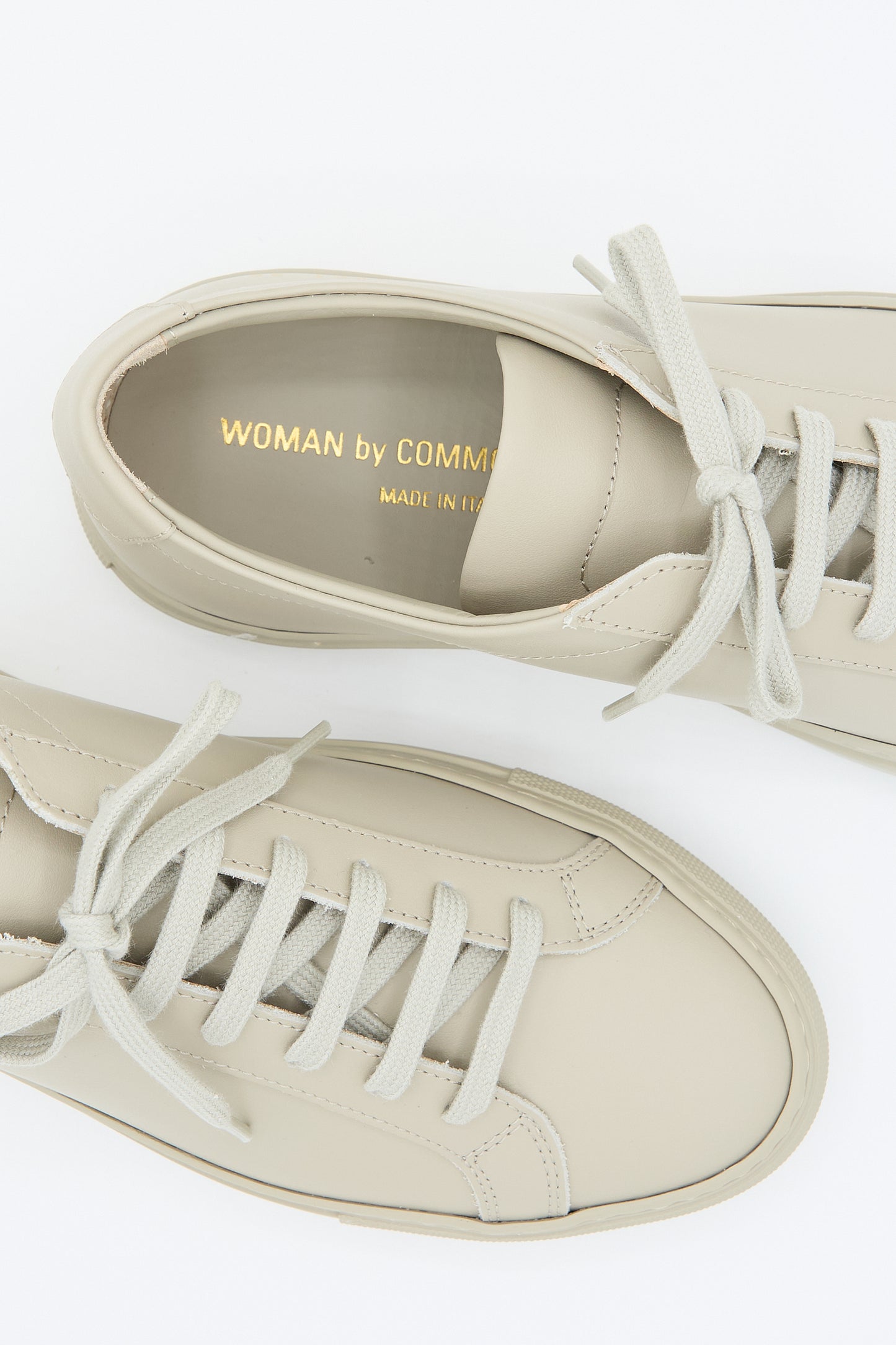 A pair of beige, minimalist Original Achilles Low 3701 sneakers with gray laces from Common Projects, isolated on a white background. Made in Italy.