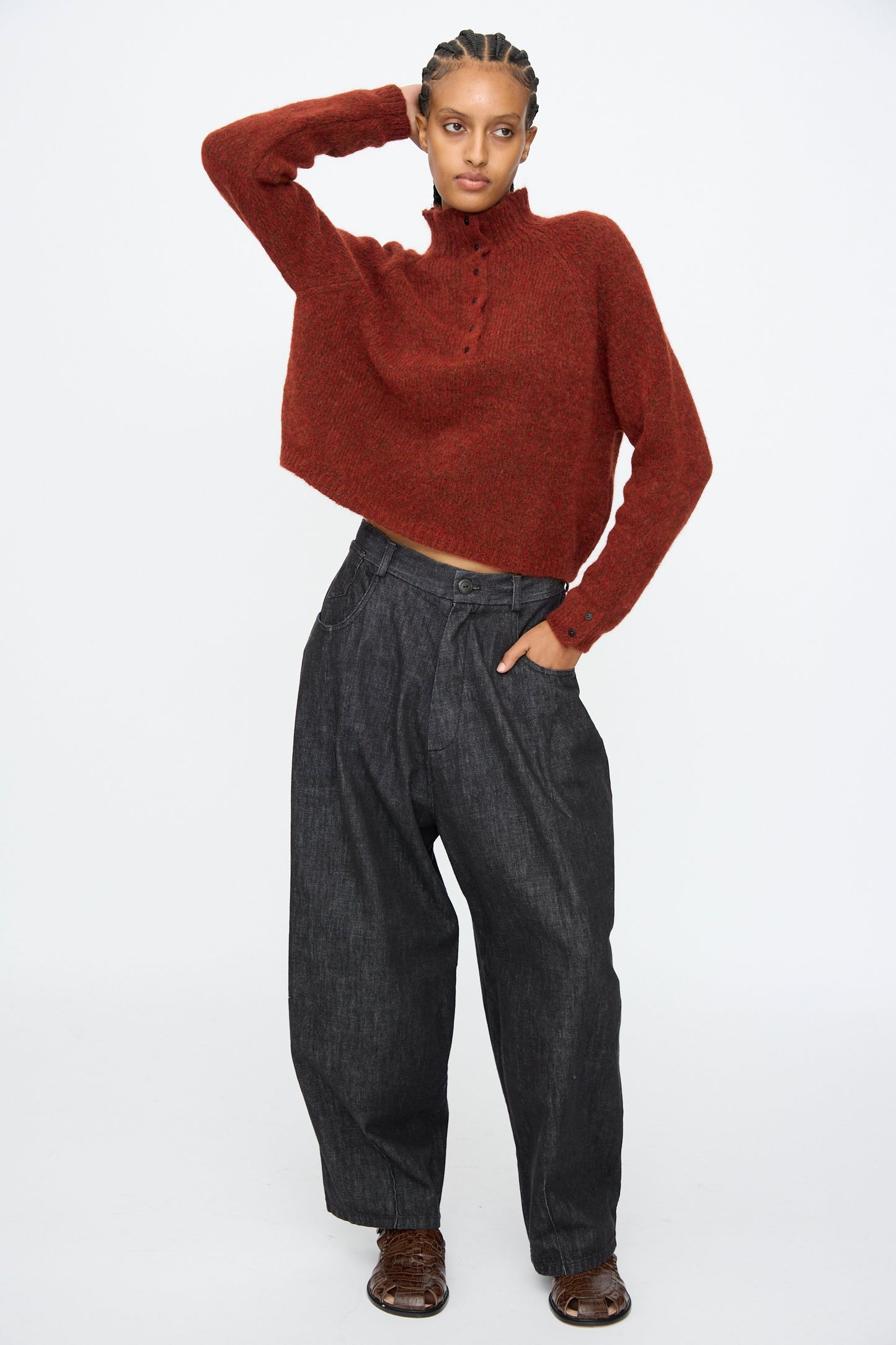 A person wears the Cordera Baby Alpaca Polo Sweater in Terracotta with loose dark gray pants, posing with one hand on their head against a plain white background.