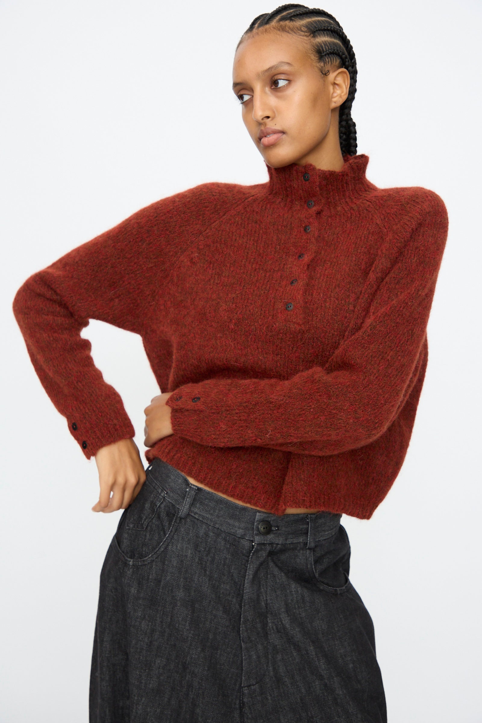 A person wears the Cordera Baby Alpaca Polo Sweater in Terracotta and dark loose-fitting pants, standing with one arm bent and gazing to the side against a plain backdrop.