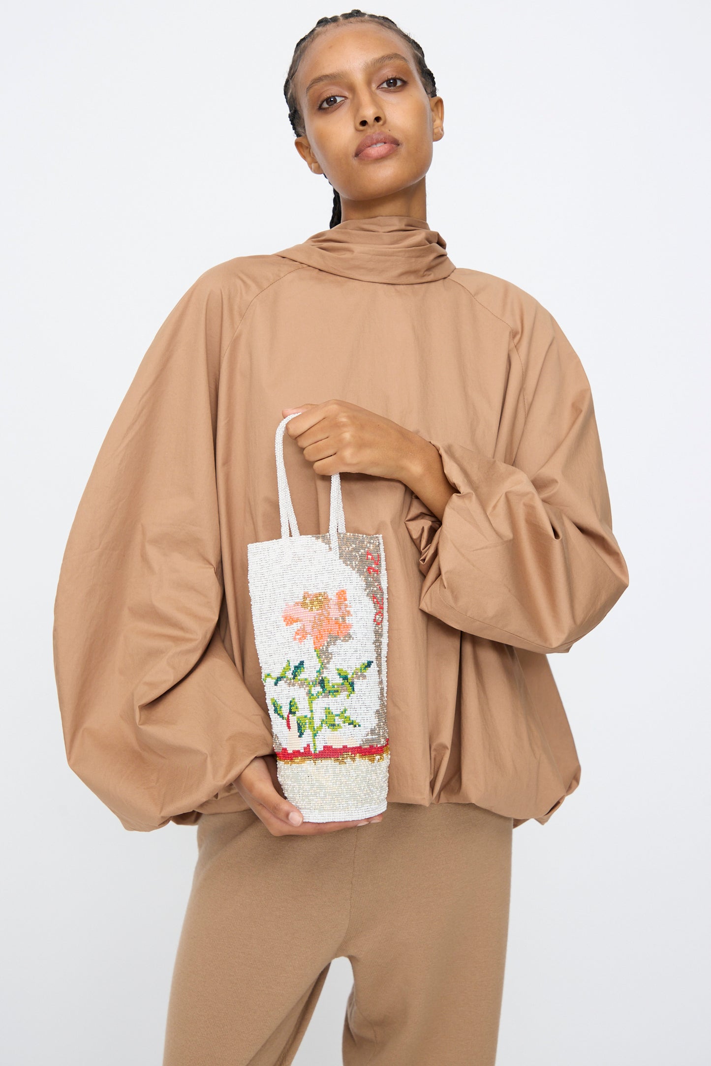 A person in loose brown clothes holds the Cordera Beaded Bag in Fiore, a white handcrafted accessory featuring a floral design inspired by Guatemalan artistry, against a plain white background.