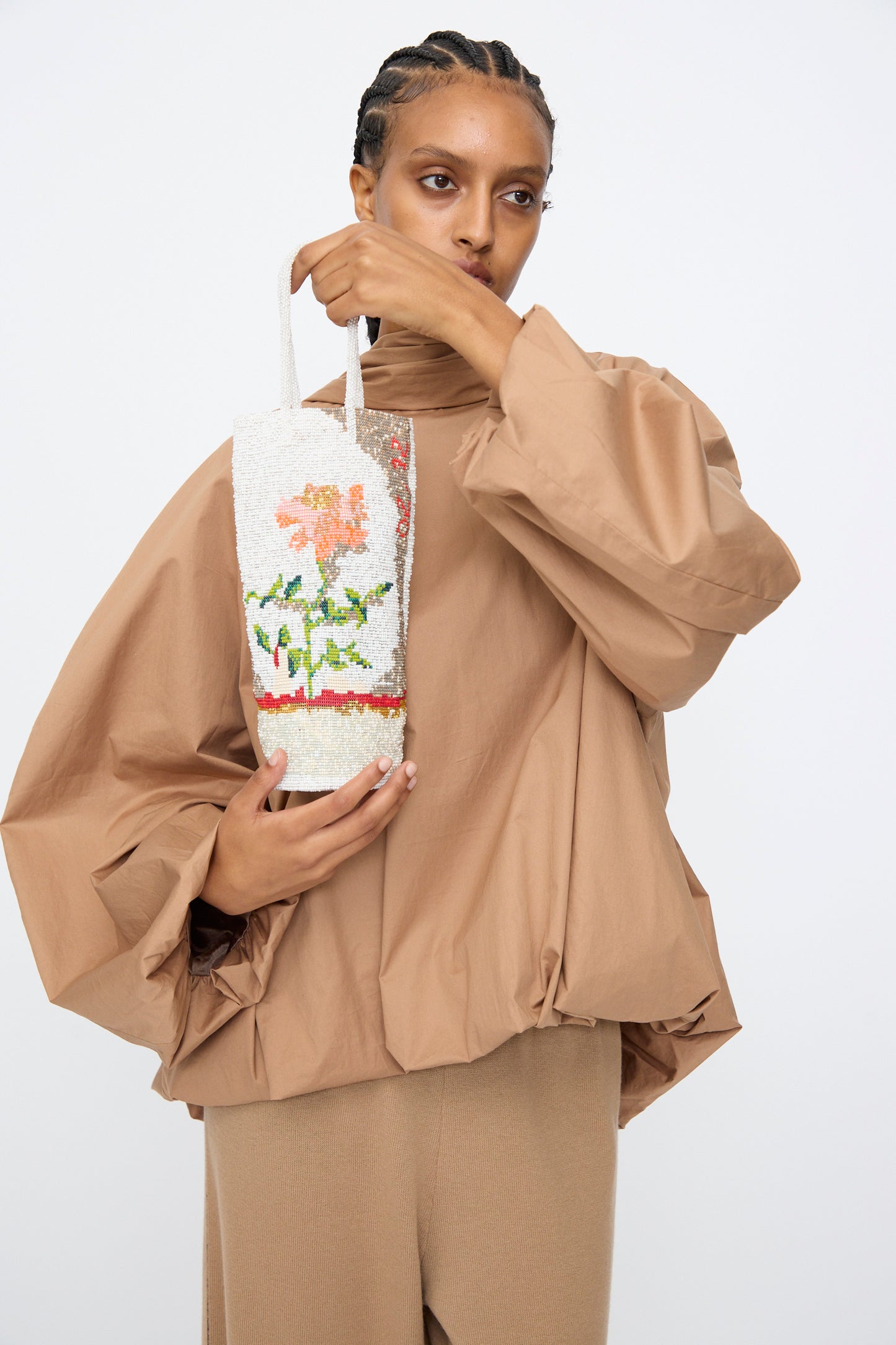 A person in a loose beige outfit holds the Cordera Beaded Bag in Fiore, featuring a handcrafted floral design inspired by Guatemala, against a plain white background.