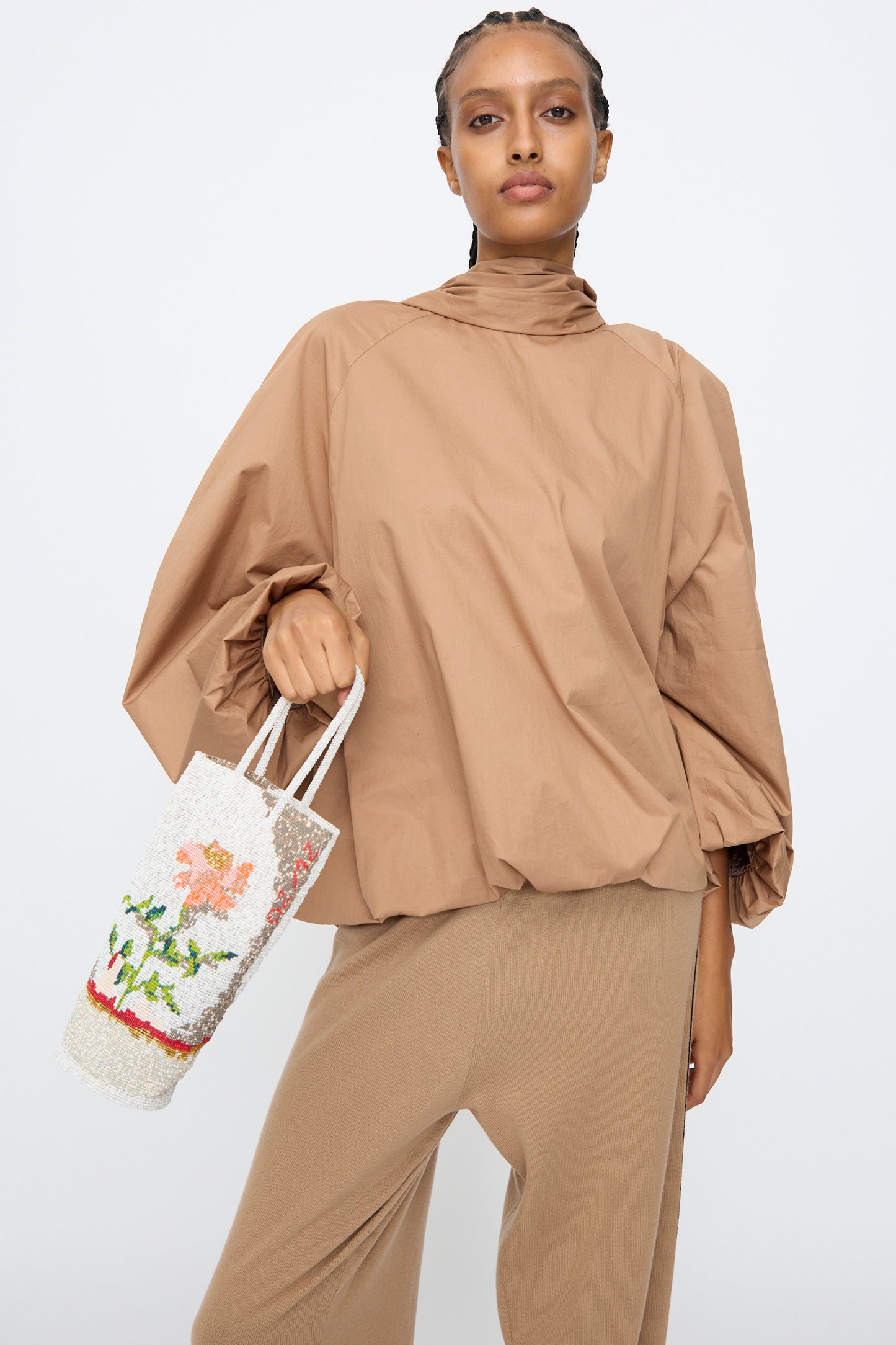 A person in a loose brown long-sleeve top and tan pants holds Cordera’s Beaded Bag in Fiore, featuring a handcrafted floral design, against a plain background.