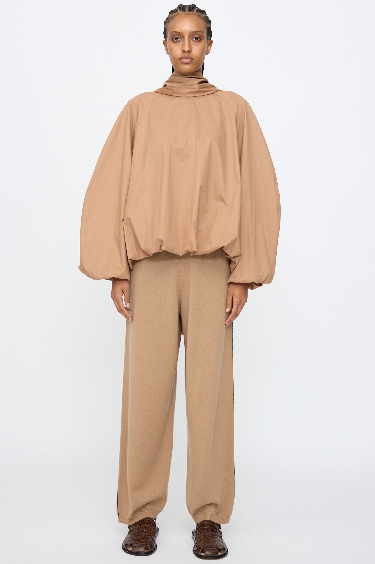 A person wears Cordera’s Bow Shirt in Camel, featuring a loose balloon silhouette with voluminous sleeves, paired with wide-leg tan pants, standing against a plain white background.