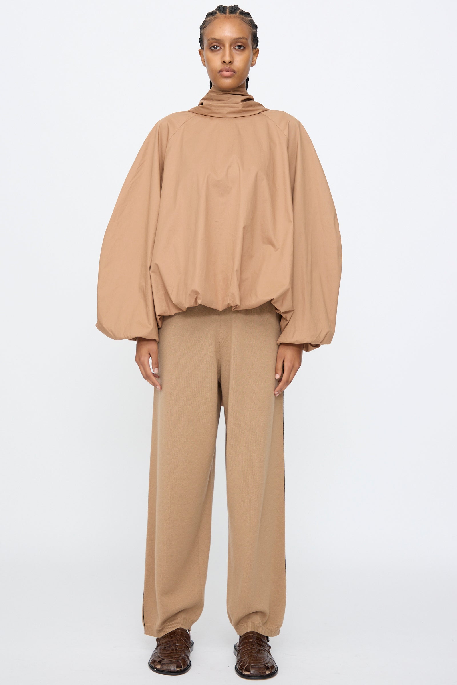 A person wears Cordera’s Bow Shirt in Camel, featuring a loose balloon silhouette with voluminous sleeves, paired with wide-leg tan pants, standing against a plain white background.