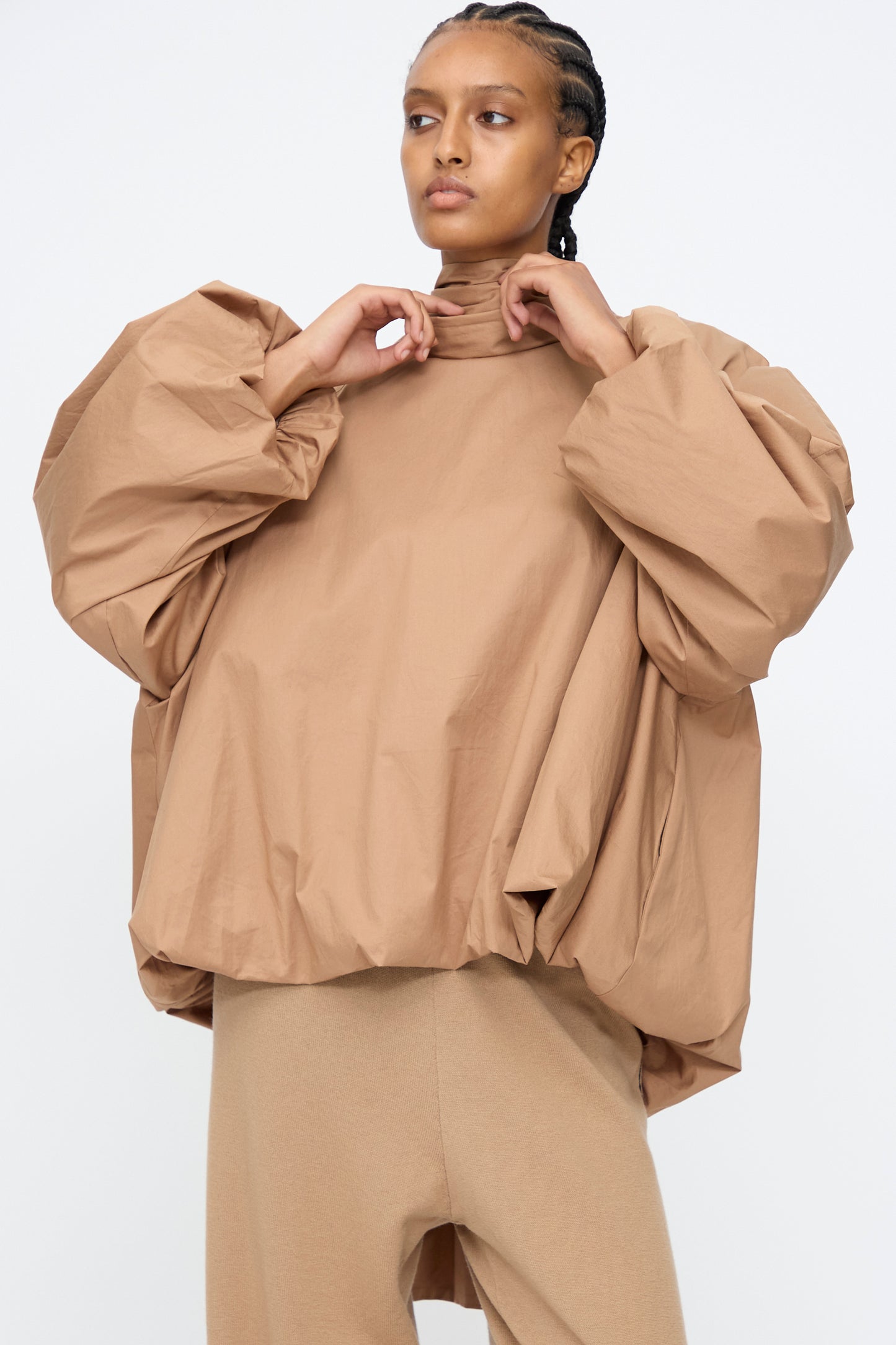 A person wears the Cordera Bow Shirt in Camel—a tan cotton blend balloon silhouette top with voluminous sleeves—paired with matching tan pants, standing against a plain white background.