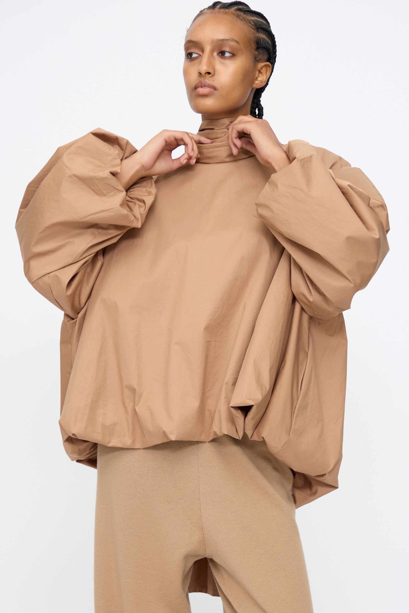 A person wears the Cordera Bow Shirt in Camel—a tan cotton blend balloon silhouette top with voluminous sleeves—paired with matching tan pants, standing against a plain white background.