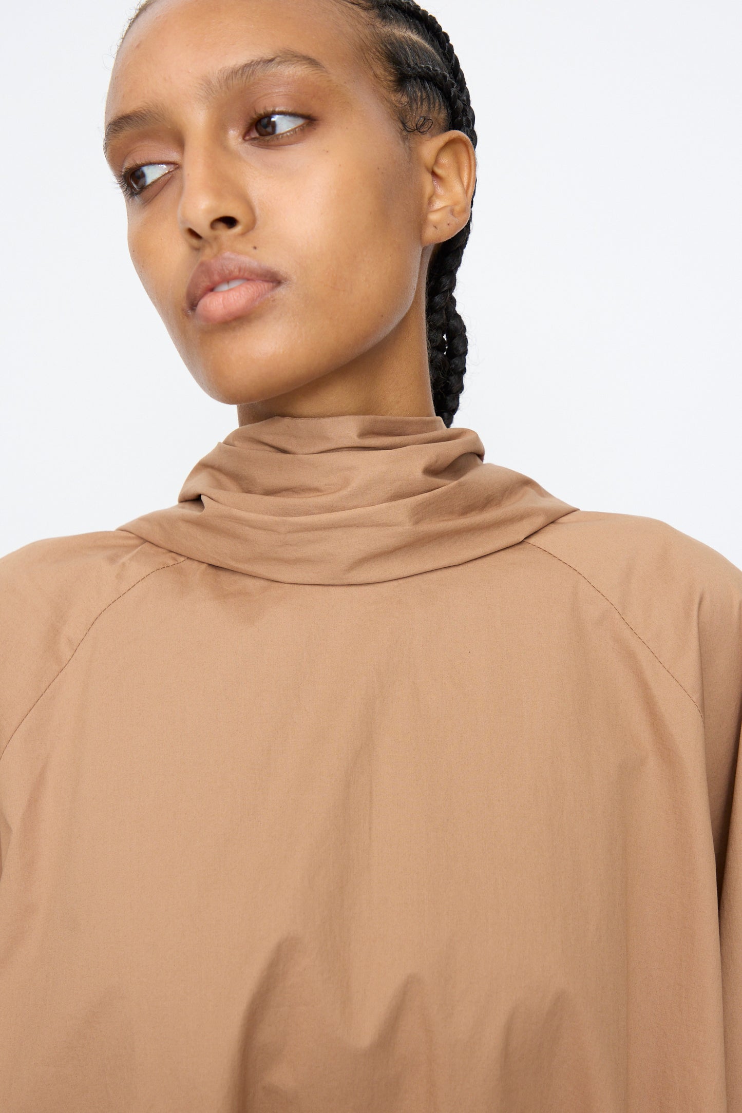 Woman with braided hair wears the Cordera Bow Shirt in Camel—a high-neck, cotton blend top with voluminous sleeves—posing against a plain background.