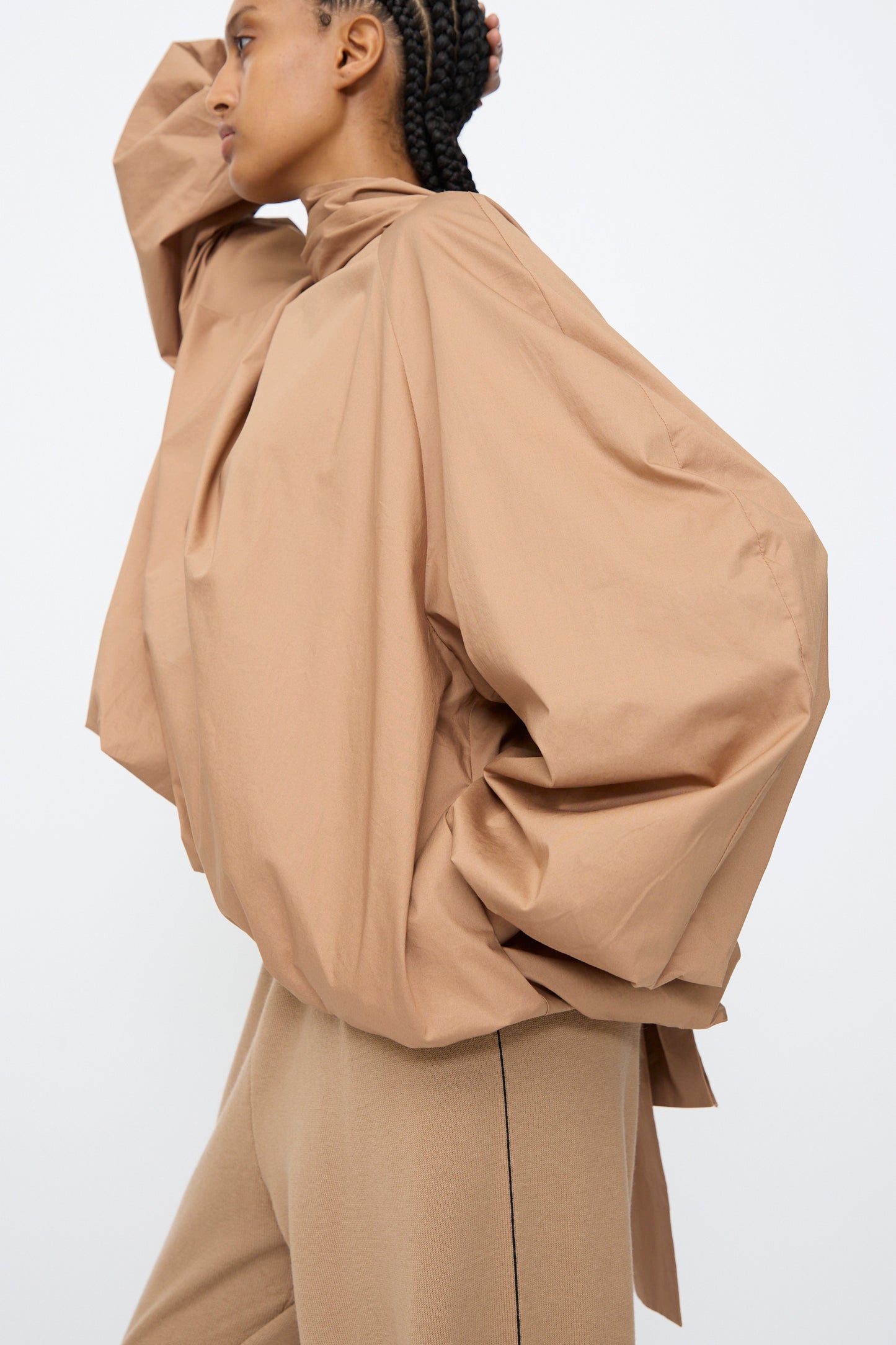Someone wears the Cordera Bow Shirt in Camel, a loose long-sleeve top, paired with matching pants, standing in profile with one arm raised to their head against a plain background.