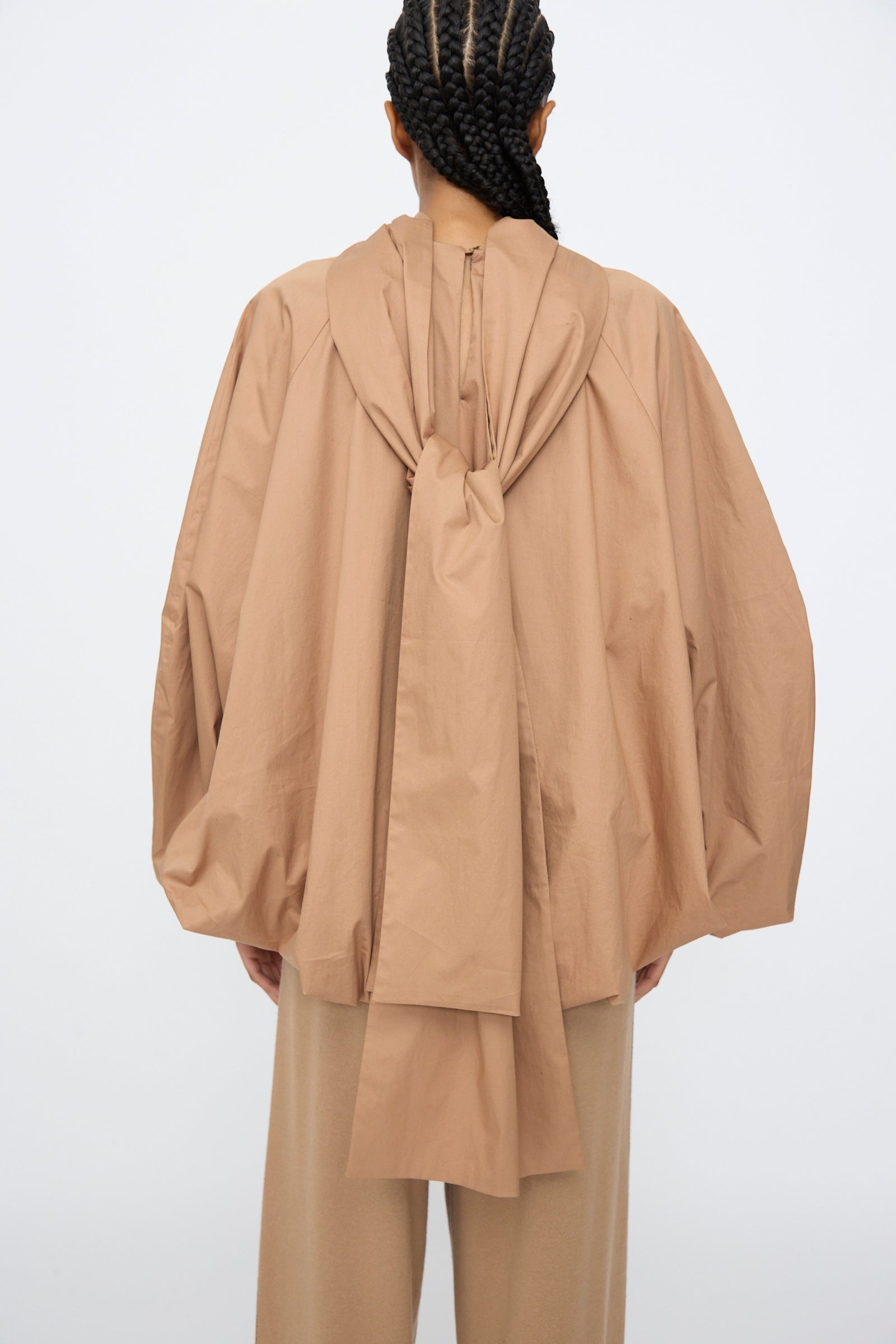 A person with braided hair models the Cordera Bow Shirt in Camel, a loose cotton blend top featuring voluminous sleeves and an oversized back bow, photographed from behind against a plain background.