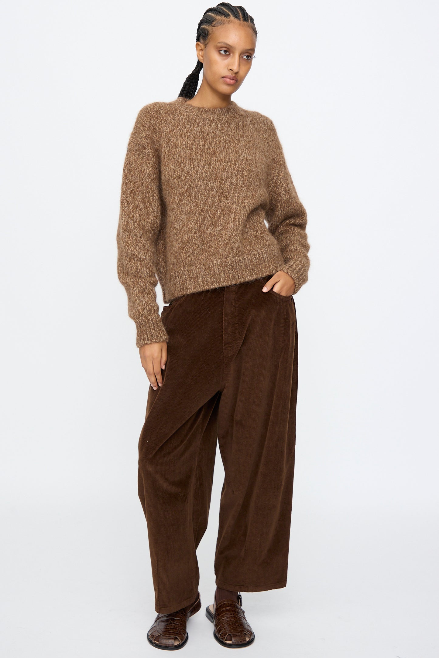 A person stands against a plain background wearing Cordera's Chunky Sweater in Brown Melange, paired with loose dark brown corduroy pants and brown shoes.