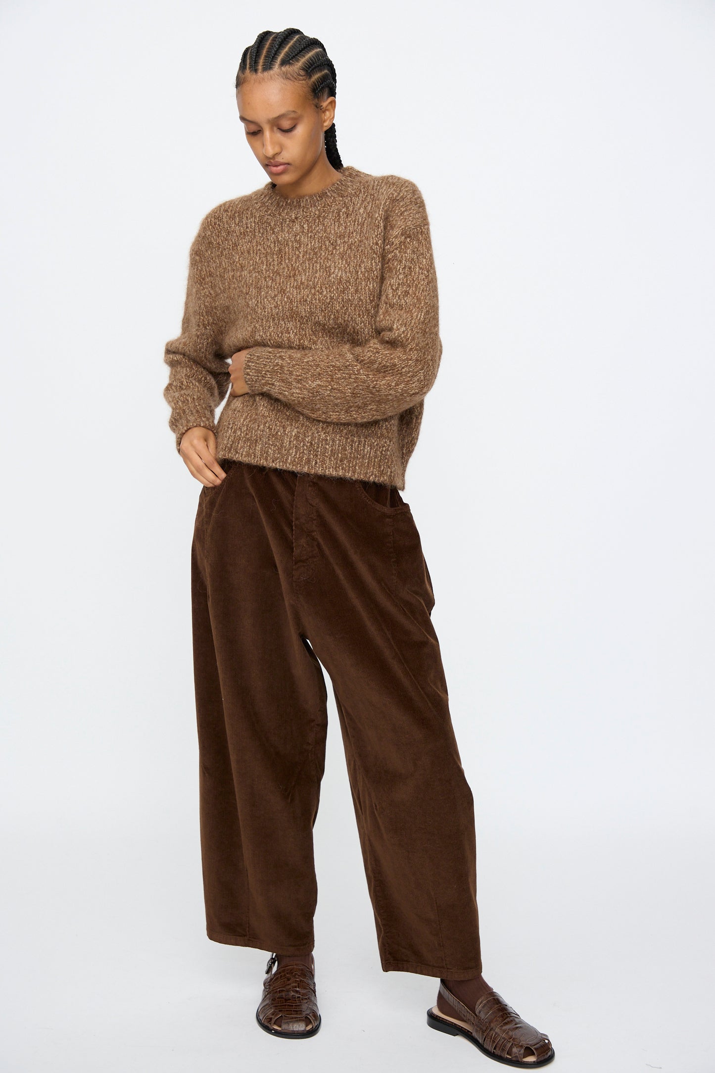 A person wears the Cordera Chunky Sweater in Brown Melange, wide brown corduroy pants, and brown sandals, standing against a plain white background.