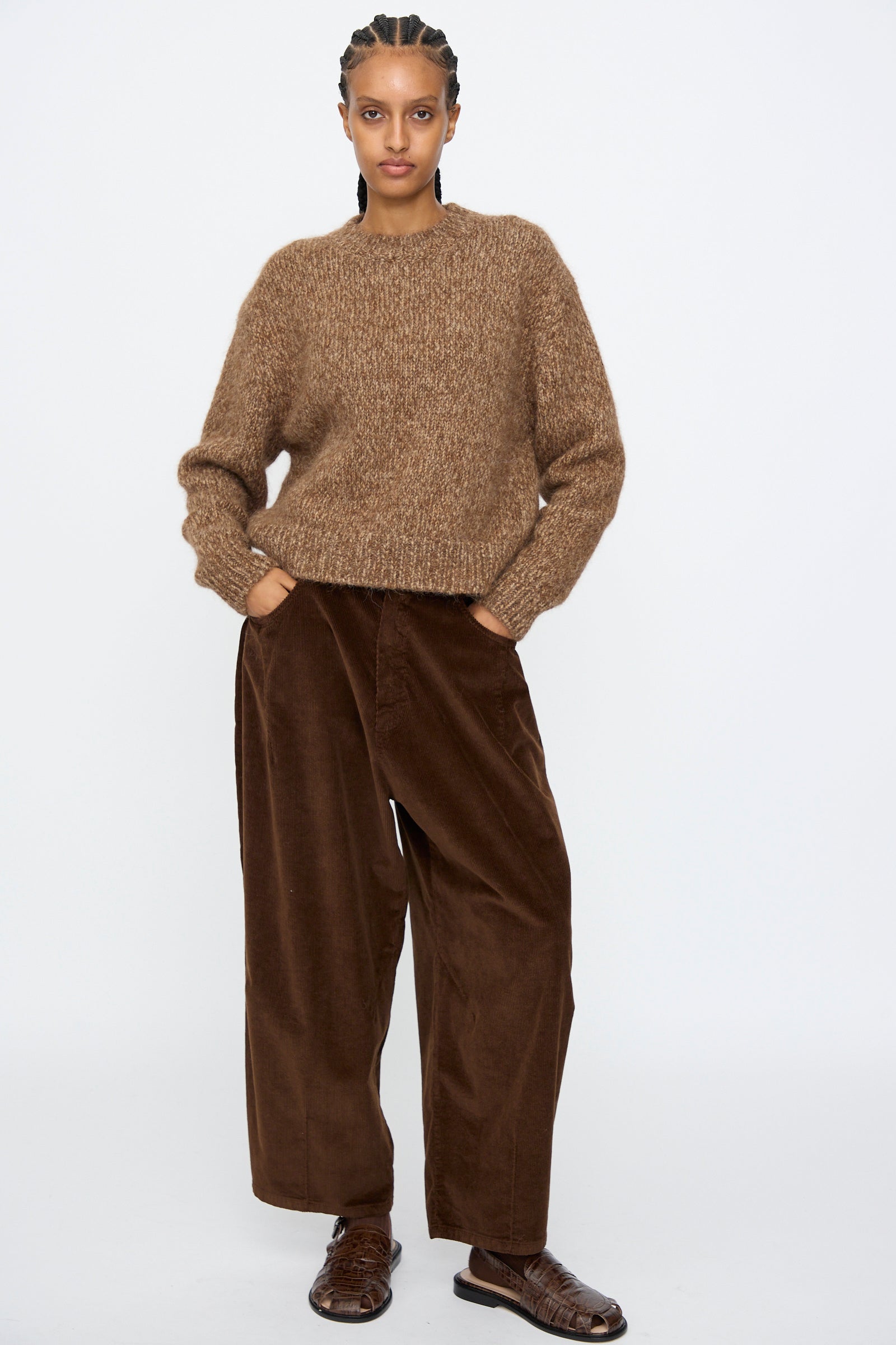 A person stands against a plain background wearing Cordera's Chunky Sweater in Brown Melange, wide-leg corduroy pants, and brown woven shoes, hands in their pockets.