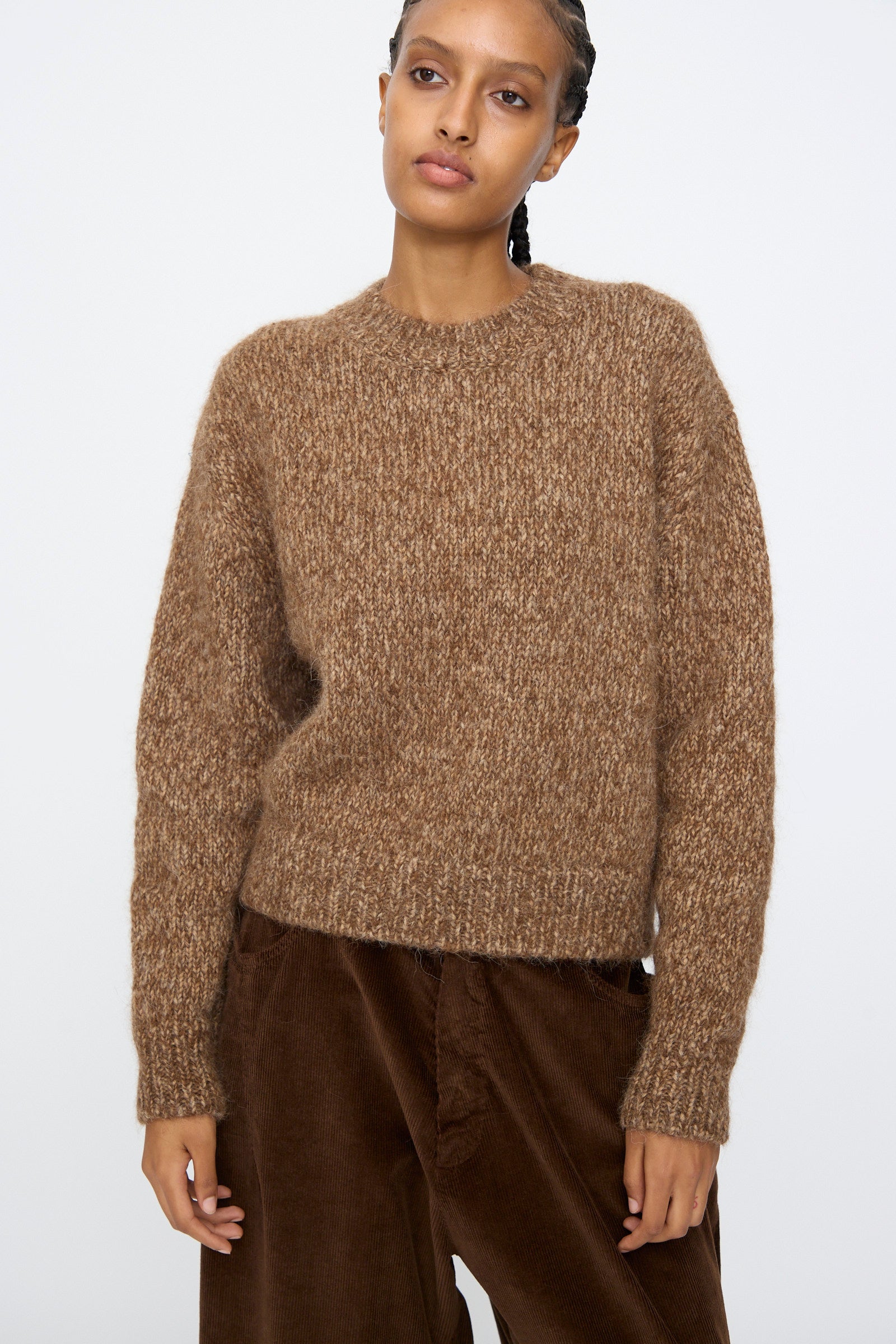 A person stands against a plain light background wearing the Cordera Chunky Sweater in Brown Melange and dark brown corduroy pants.