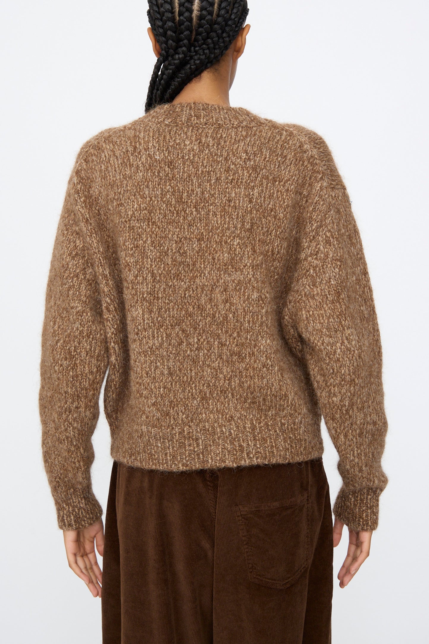 A person with braided hair is seen from the back, wearing the Cordera Chunky Sweater in Brown Melange and brown corduroy pants, posed against a plain background.