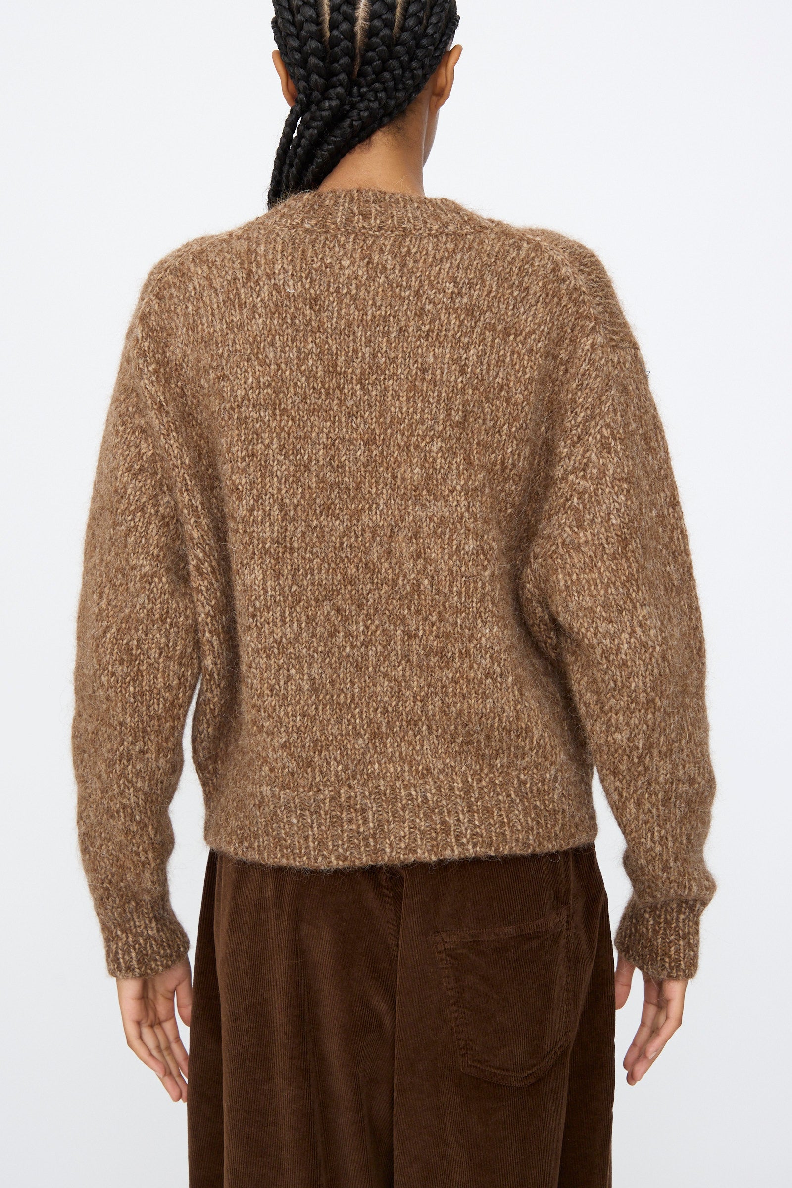 A person with braided hair is seen from the back, wearing the Cordera Chunky Sweater in Brown Melange and brown corduroy pants, posed against a plain background.