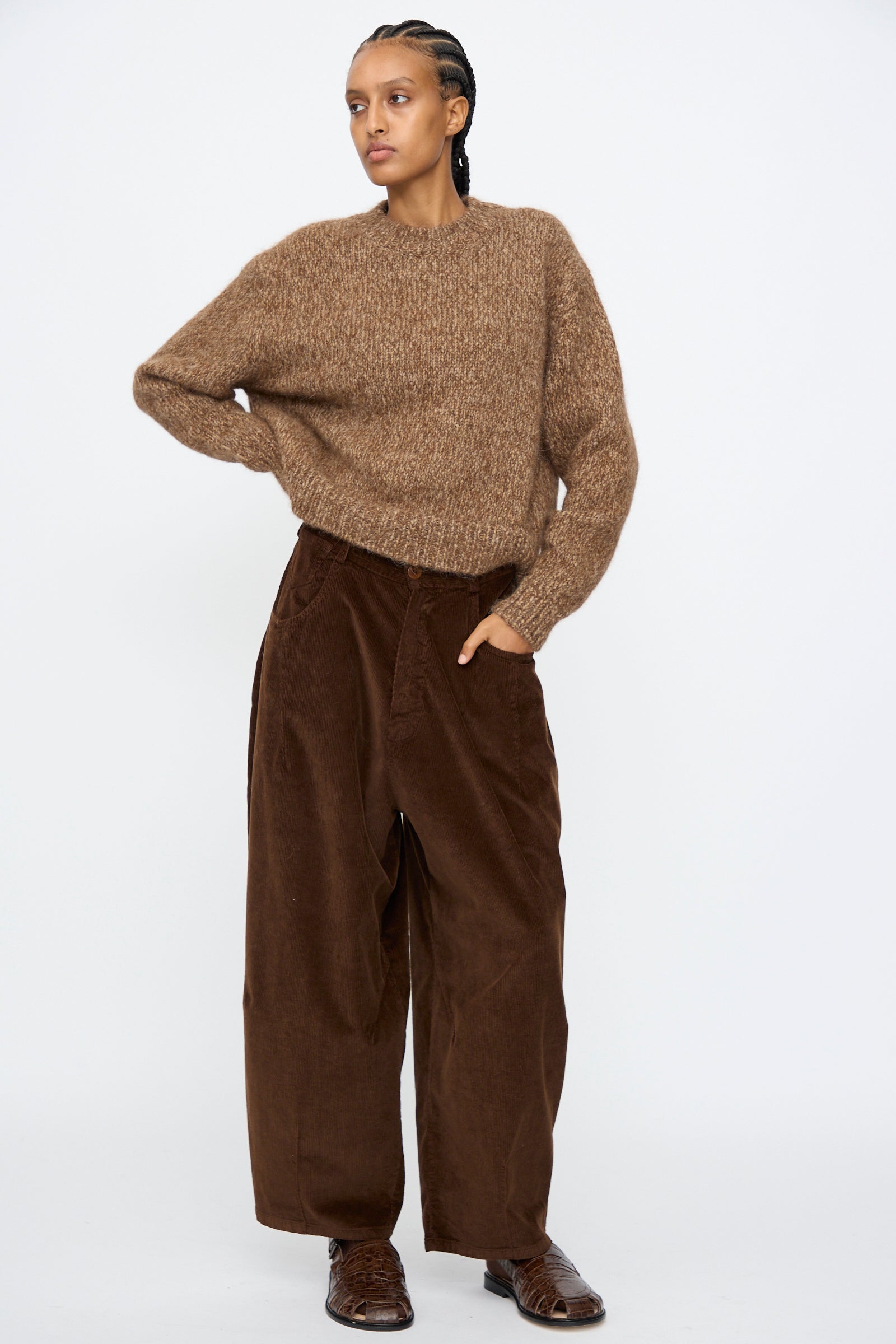 A person stands against a plain white background, wearing Cordera’s Corduroy Baggy Pants in Brown, paired with a brown knit sweater and brown shoes.