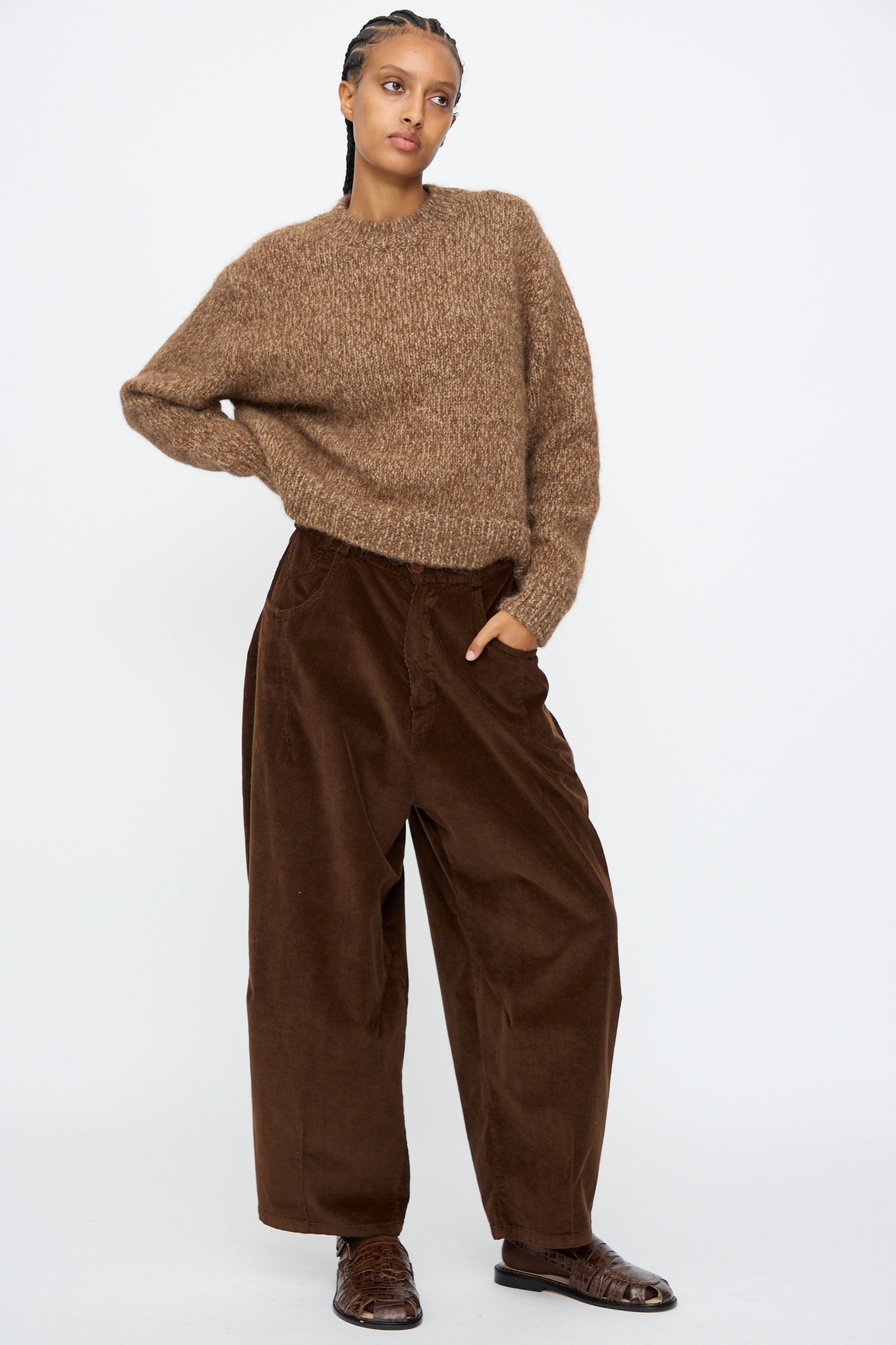 A person stands against a plain background wearing Cordera's Corduroy Baggy Pants in Brown, featuring a wide-leg fit and elastic waistband, paired with a brown textured sweater and brown shoes.