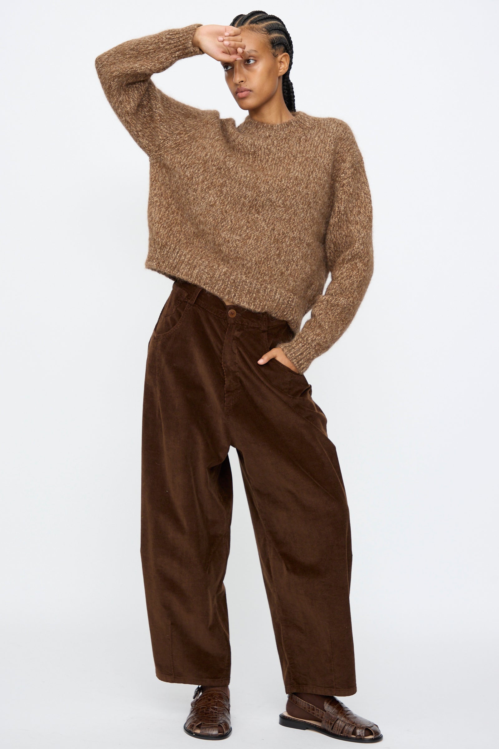 A person stands against a plain background, one hand on hip and the other shading their eyes, wearing Cordera’s Corduroy Baggy Pants in Brown with an elastic waistband, paired with a loose brown sweater and brown shoes.