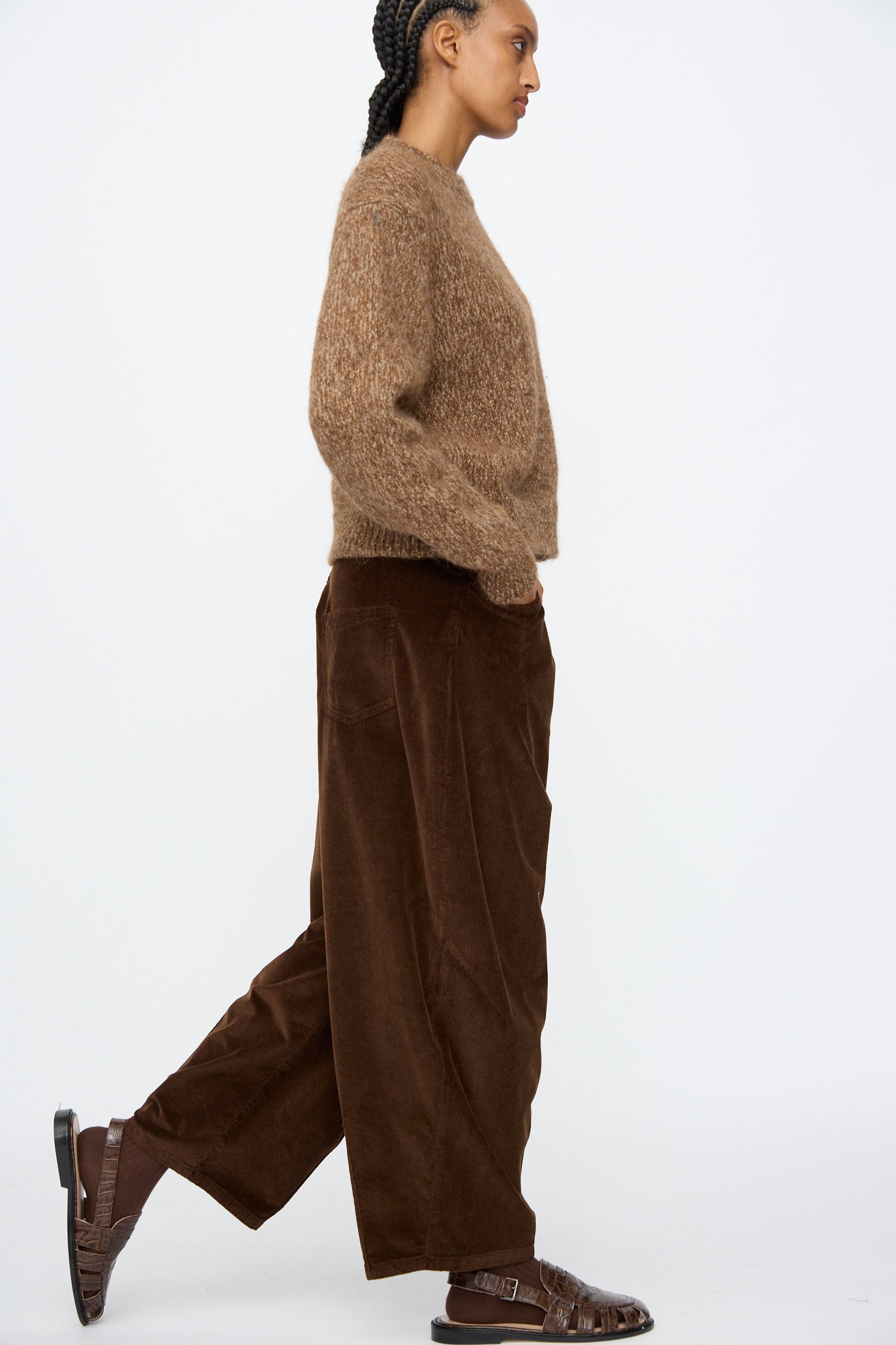 A person wearing Cordera's Corduroy Baggy Pants in Brown walks against a plain white background, styled with a brown fuzzy sweater and brown sandals.