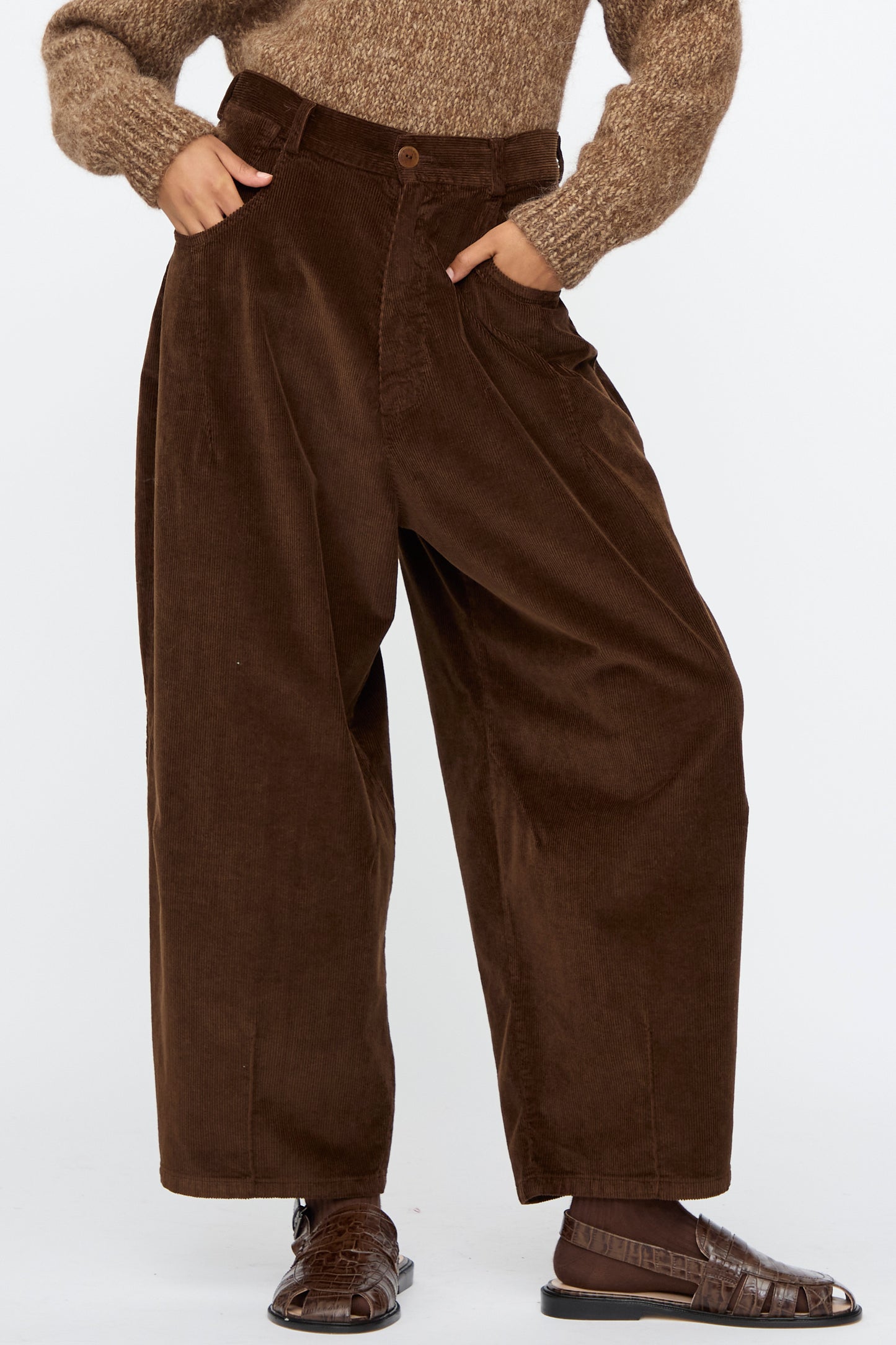 A person stands with hands in pockets against a plain background, wearing Cordera's Corduroy Baggy Pants in Brown paired with a brown sweater and brown sandals.