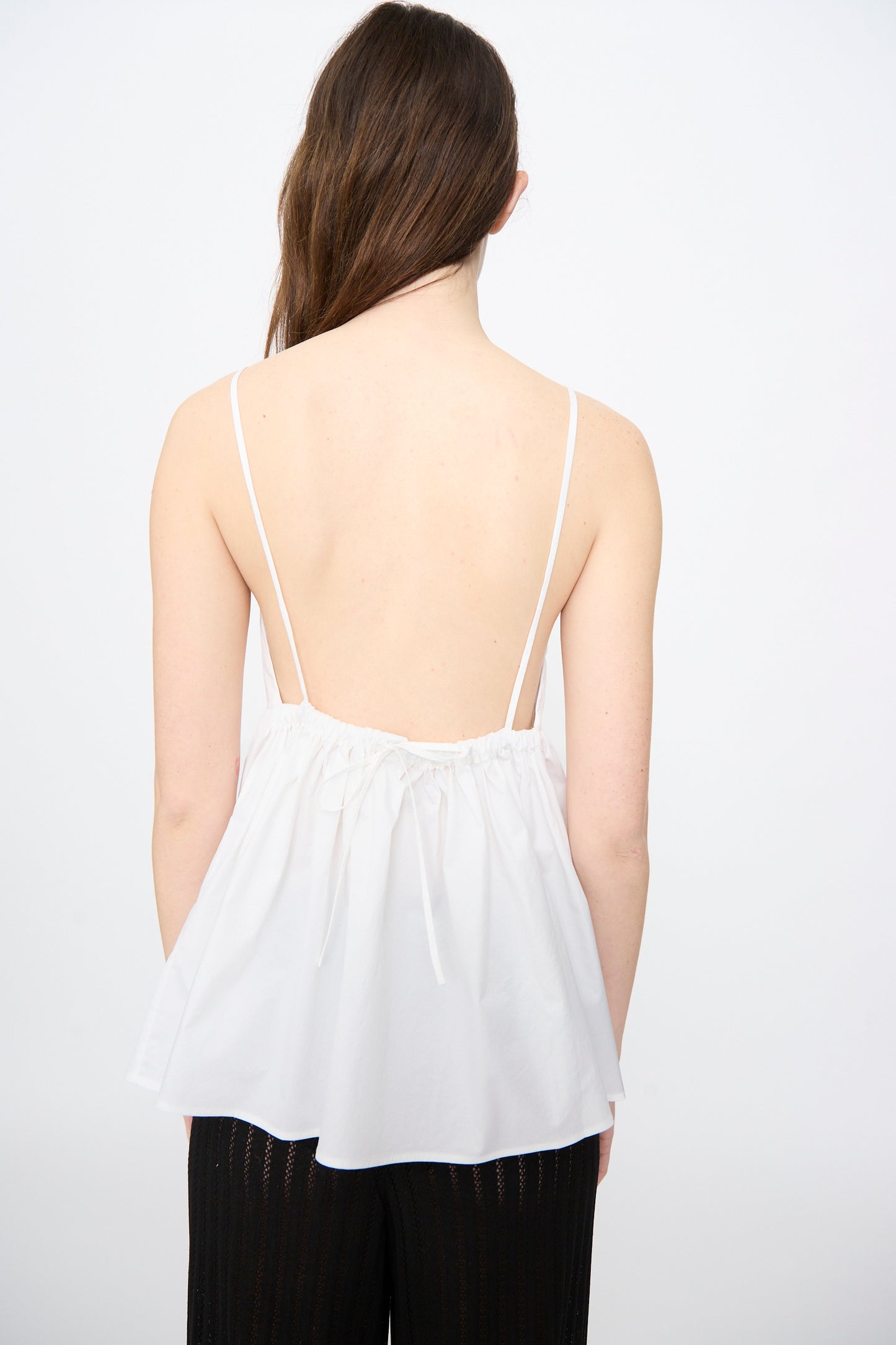 A person with long brown hair, seen from the back, wears Cordera's Cotton Embroidered Top in White with an adjustable back tie and black pants against a plain white background.