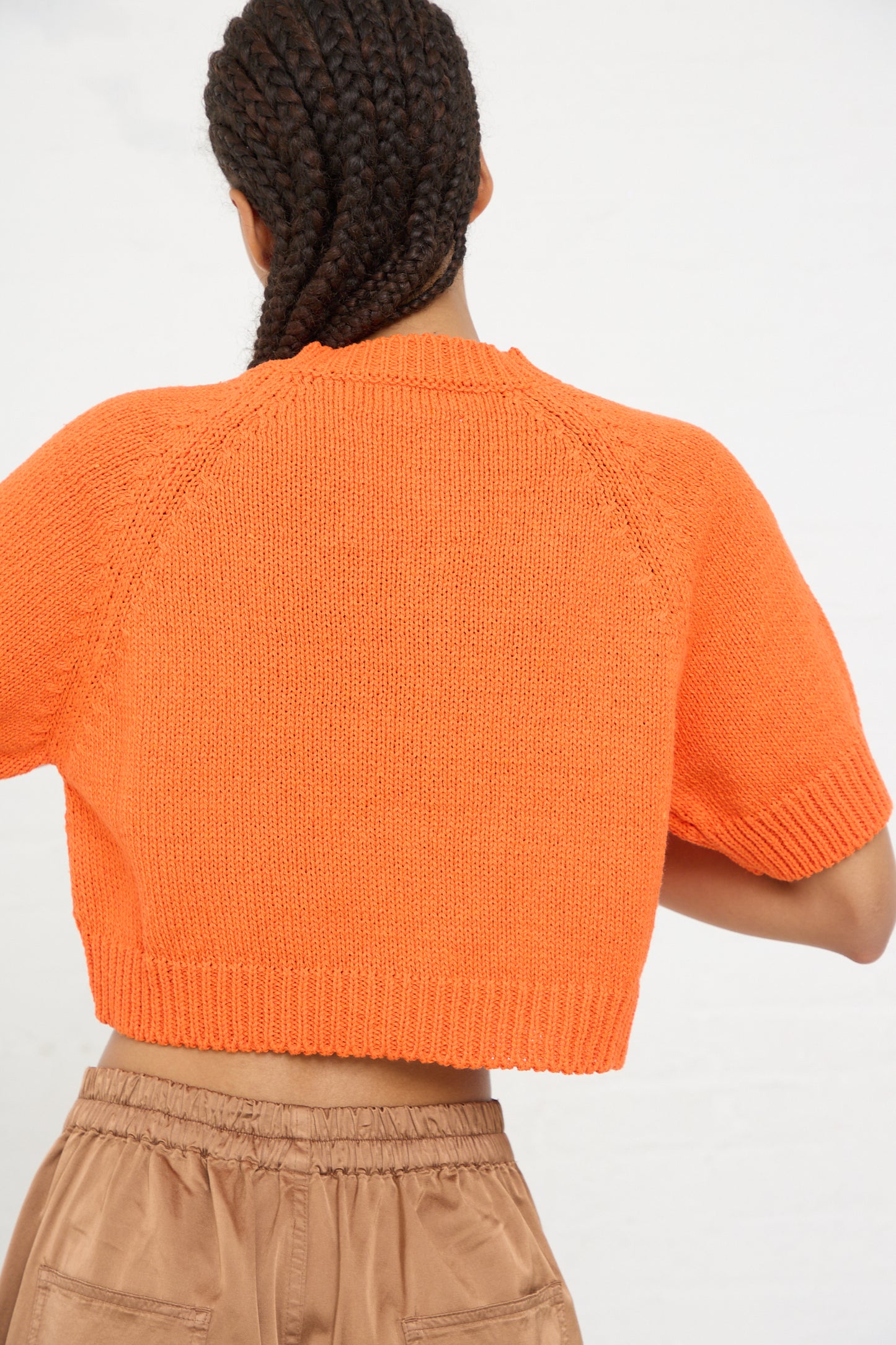 Woman from behind wearing a bright orange cropped, Cotton Knit Buttoned Top in Mandarina with beige trousers, sustainably made in Spain by Cordera.