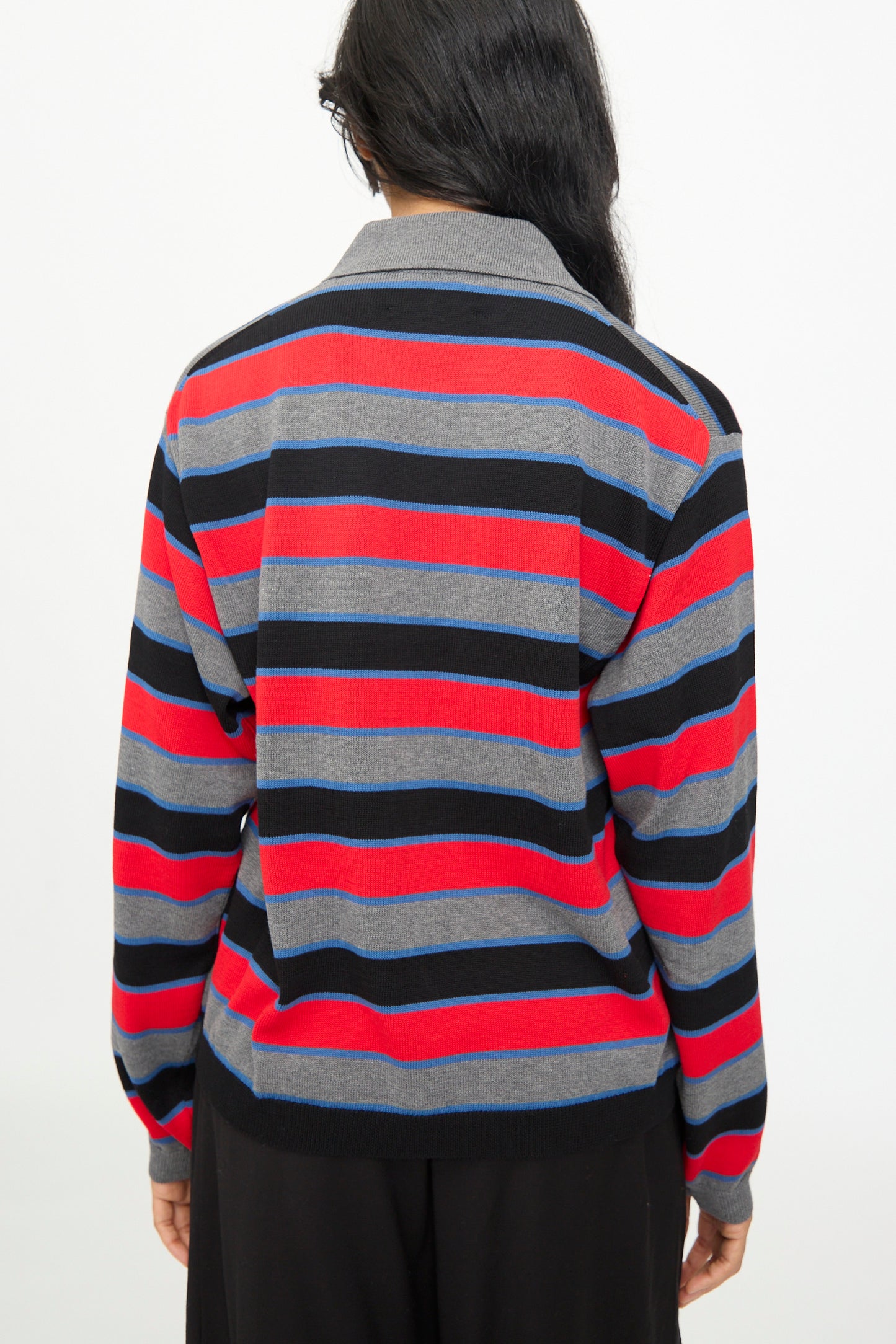 A person with long dark hair is seen from the back wearing the Cordera Cotton Long Sleeve Polo in Stripe—an oversized-fit sweater featuring gray, black, red, and blue horizontal stripes—against a plain white background.