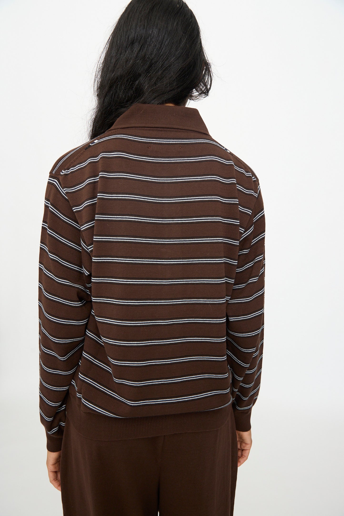 A person stands with their back to the camera, wearing the Cordera Cotton Polo Cardigan in Striped—a brown organic cotton long-sleeve shirt with white horizontal stripes—paired with matching brown pants against a plain background.
