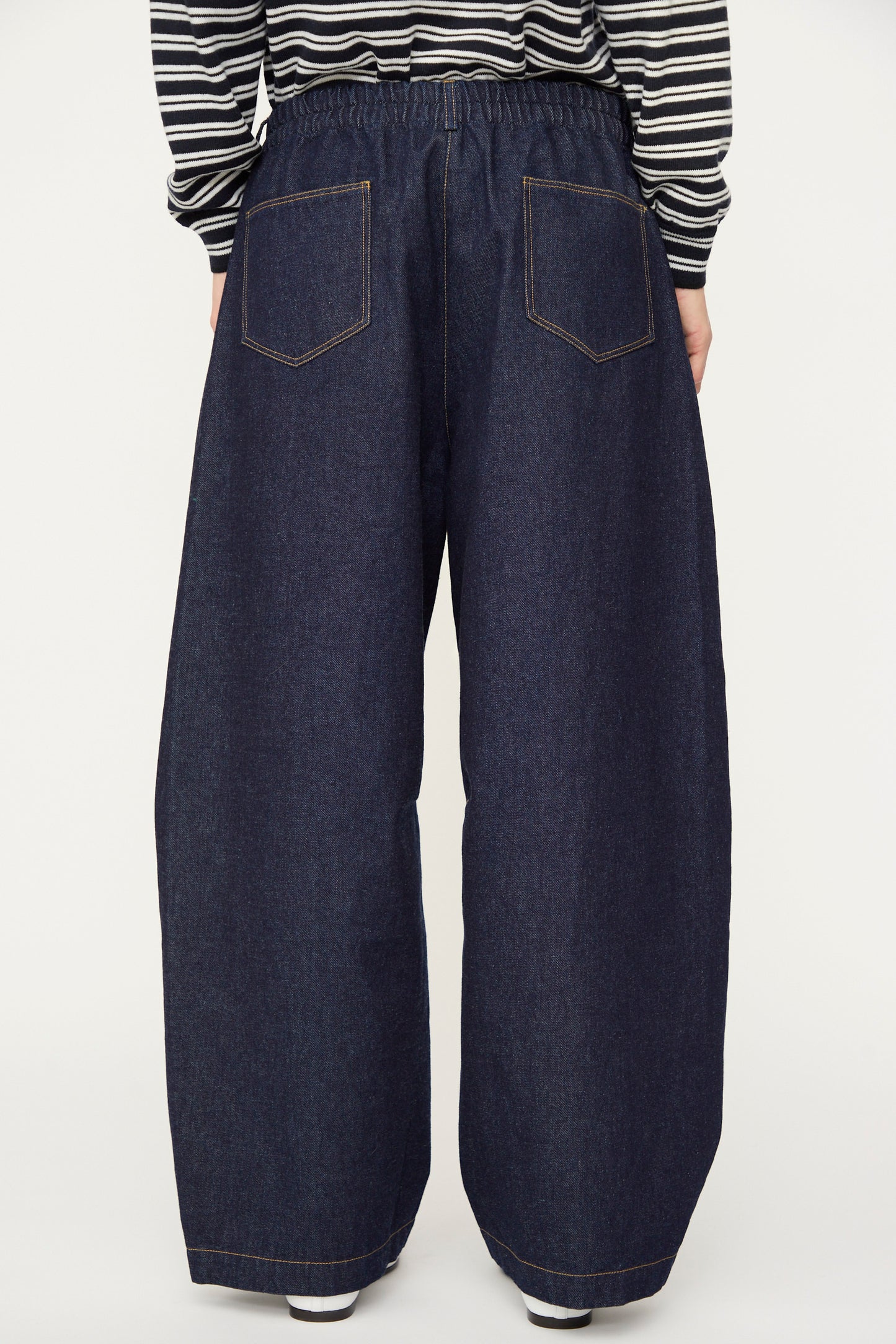 A person is shown from the back wearing Cordera’s Curved Baggy Jean in dark denim and a black-and-white striped long-sleeve shirt. The high-rise, wide-leg jeans are crafted from organic cotton.