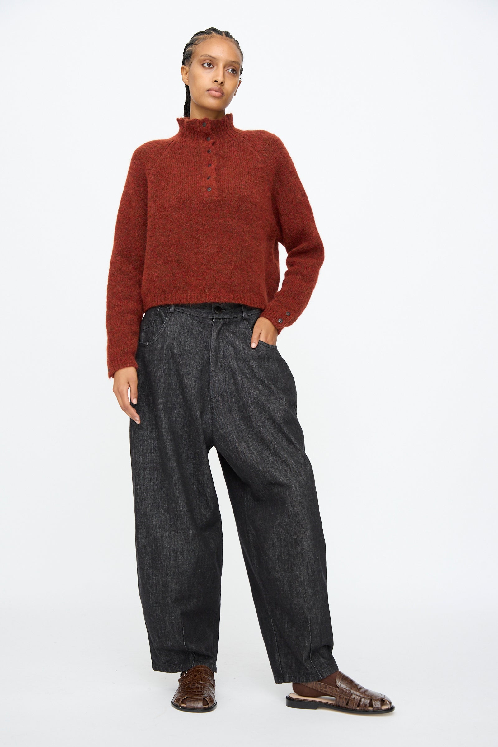 Person standing against a plain background, wearing a rust-colored sweater, Cordera Denim Baggy Pants in Black, and brown flat shoes.