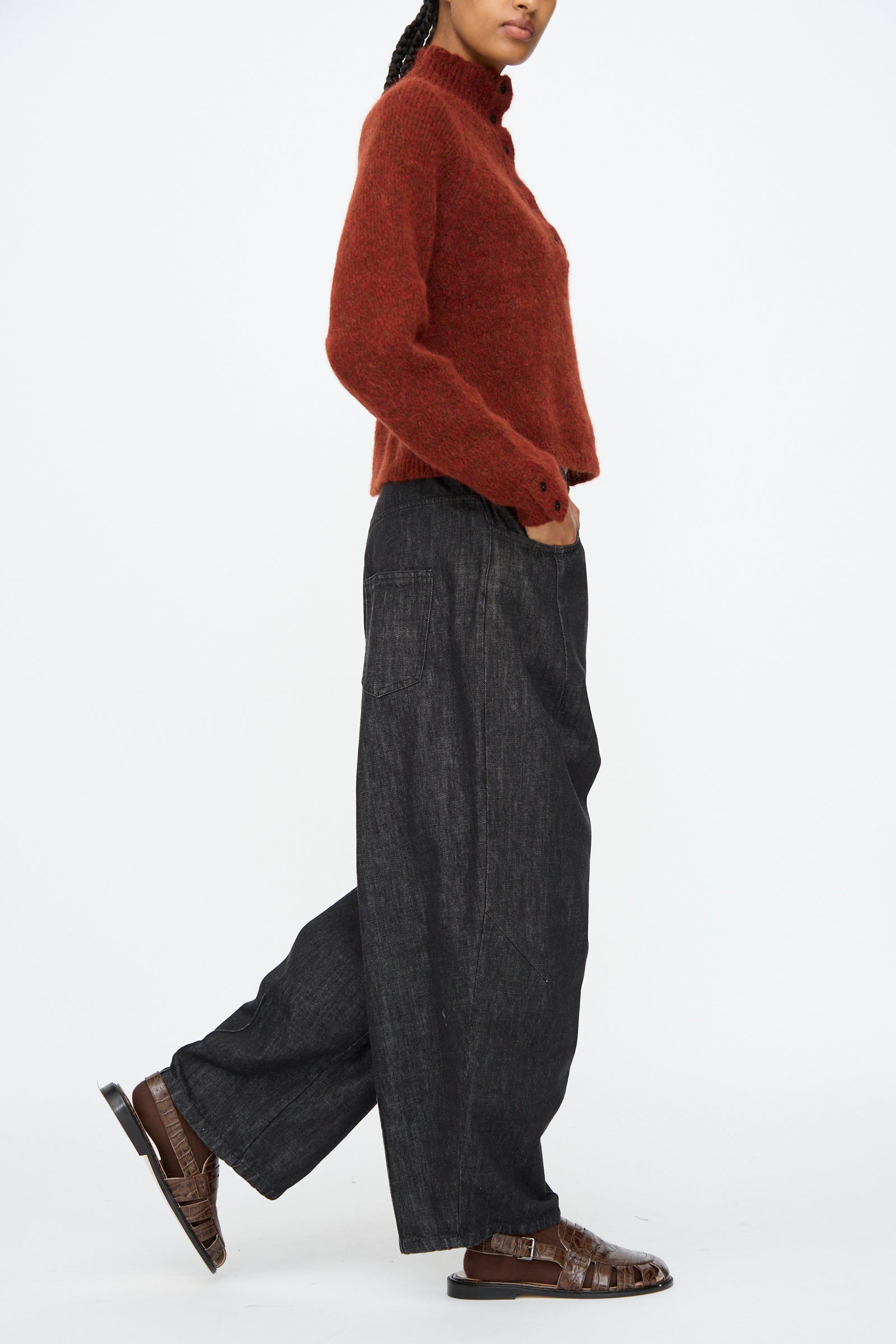 A person wearing a rust-colored sweater, Cordera Denim Baggy Pants in Black, and brown sandals walks against a plain white background.