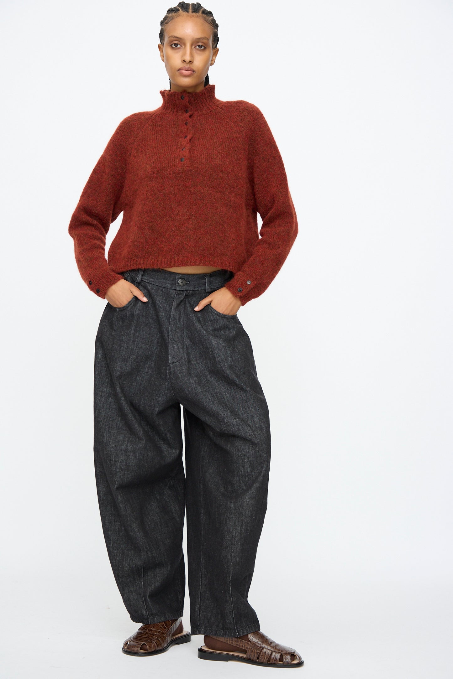 A person stands against a white background wearing a red knit sweater, Cordera Denim Baggy Pants in Black, and brown shoes, with hands in pockets.