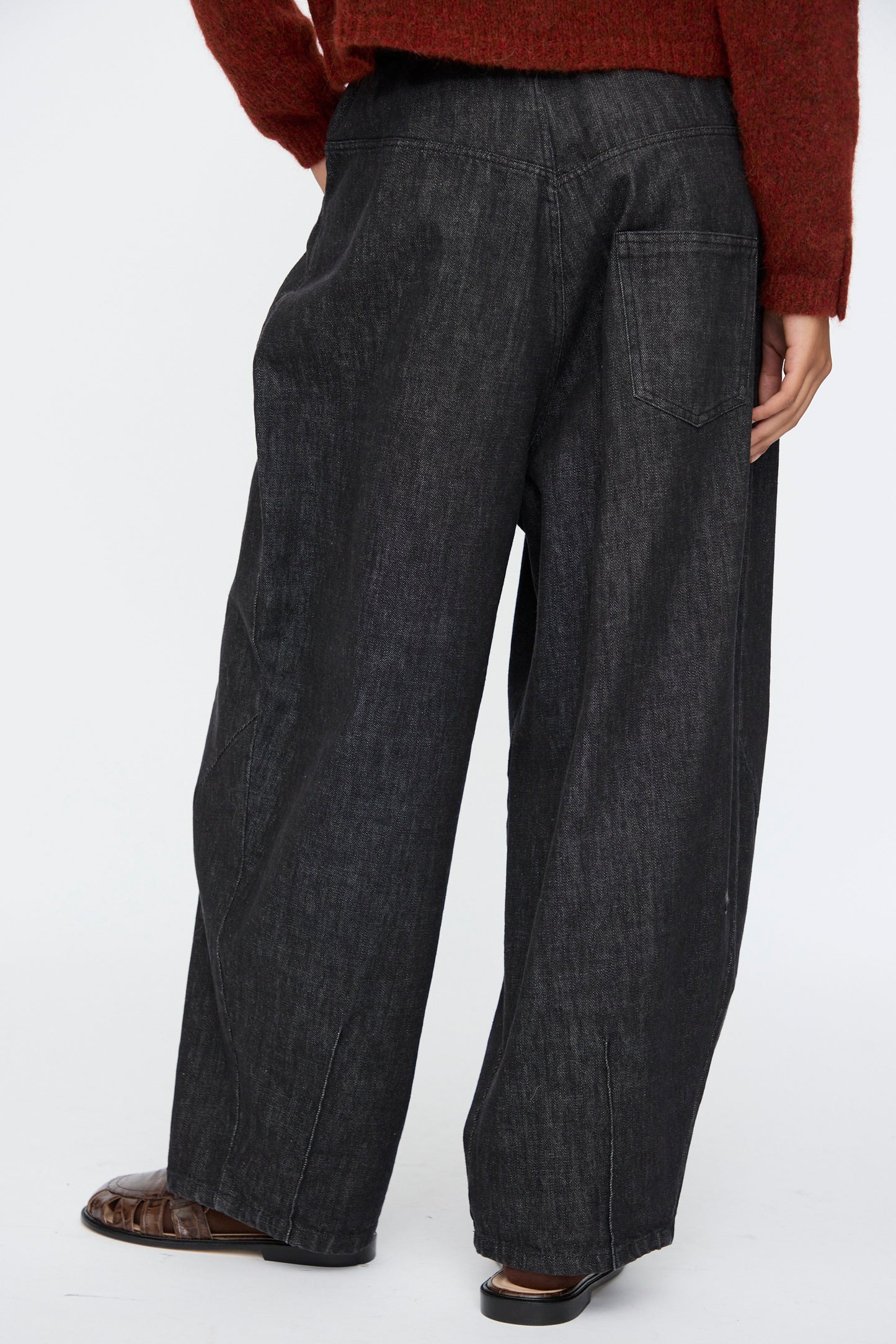 A person is shown from the back against a plain background, wearing Cordera’s Denim Baggy Pants in Black, paired with a rust-colored sweater and brown shoes.