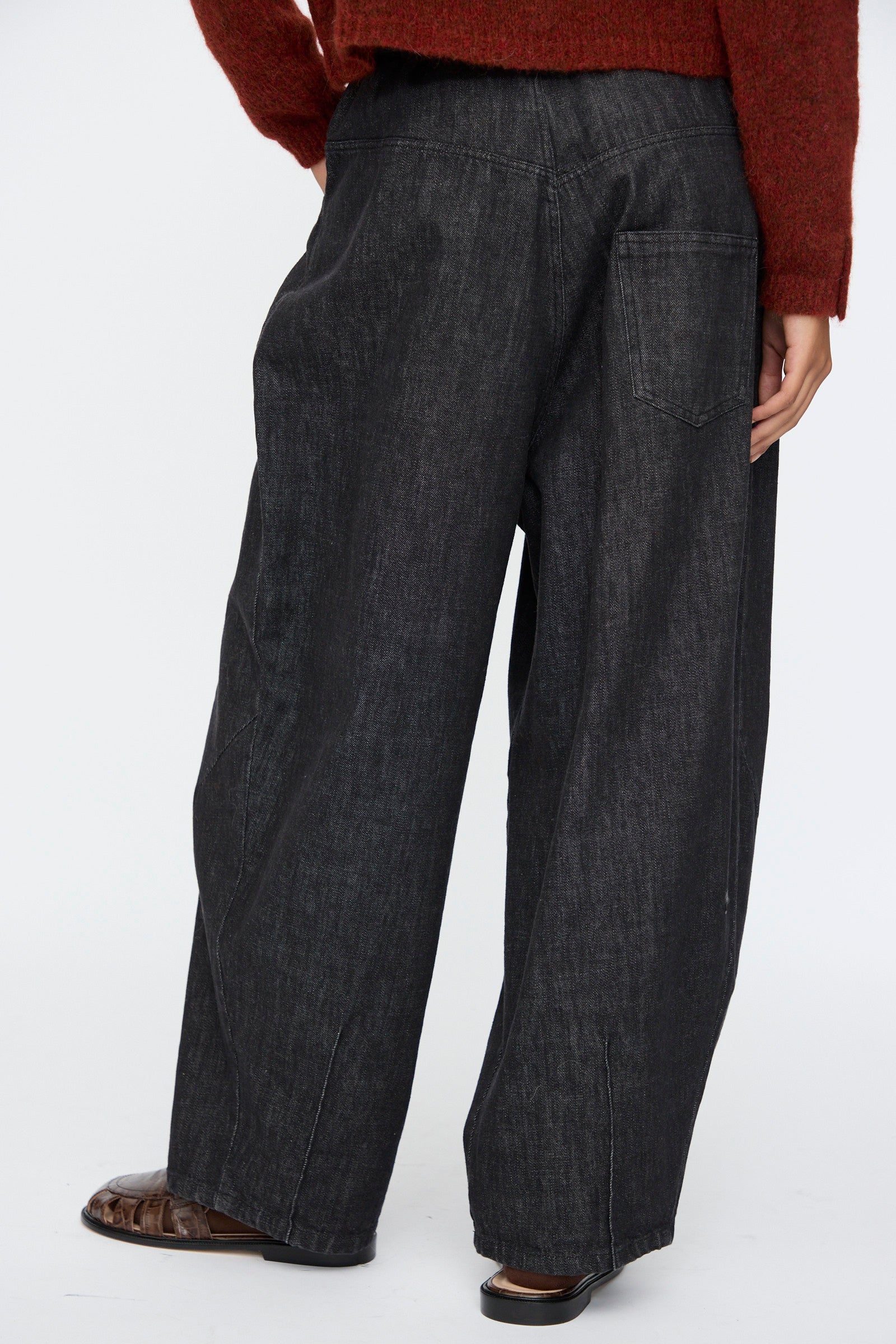 A person is shown from the back against a plain background, wearing Cordera’s Denim Baggy Pants in Black, paired with a rust-colored sweater and brown shoes.