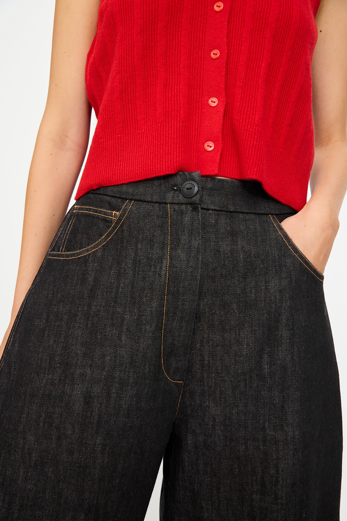 A person wears Cordera's Denim Curved Pant in Black, paired with a red sleeveless top with buttons; one hand rests in a pocket of the high-waisted organic cotton jeans.