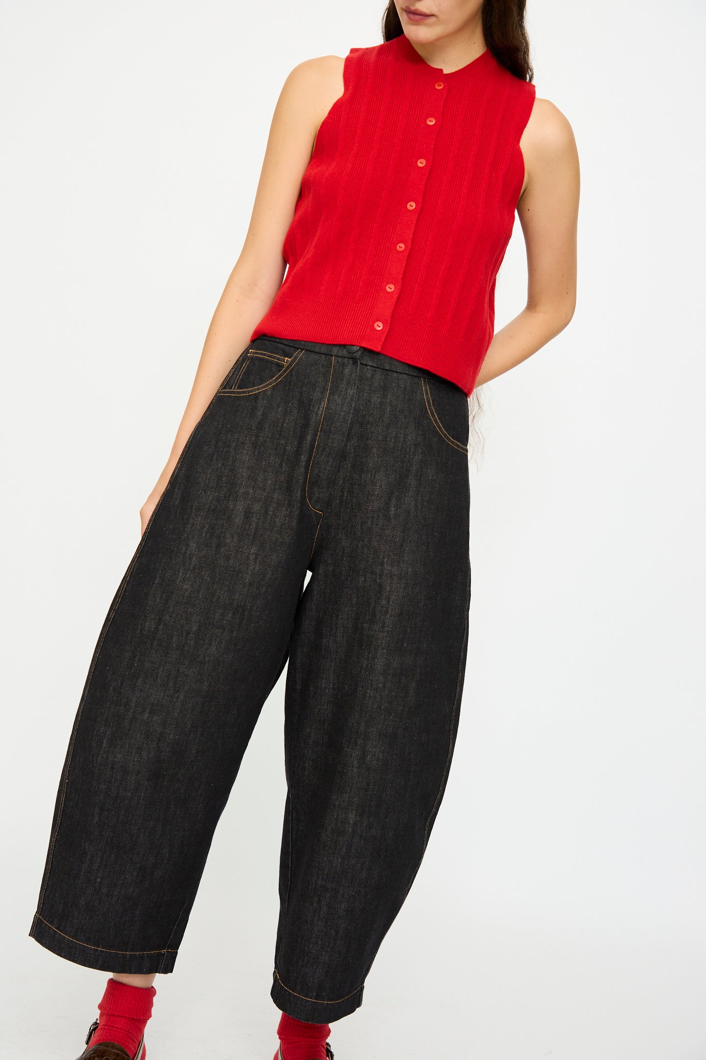 A person wears the Cordera Denim Curved Pant in Black and a sleeveless red button-up top, standing against a plain white background.