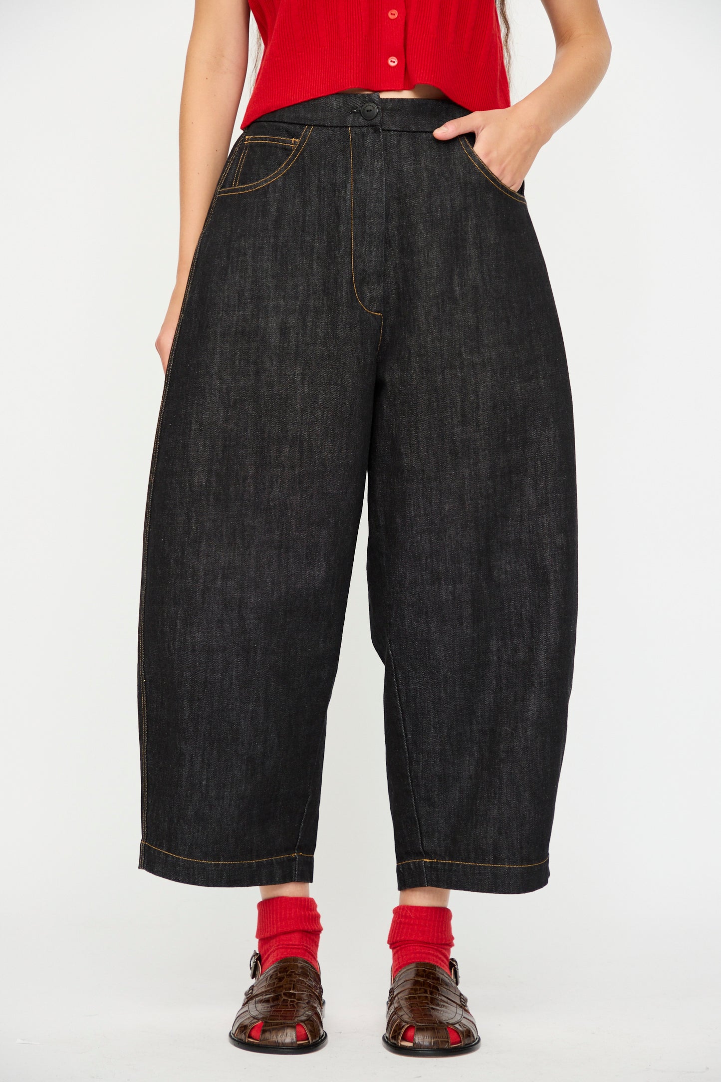 A person wears the Cordera Denim Curved Pant in Black with a red sleeveless top, red socks, and brown open-toe sandals, standing against a plain white background.