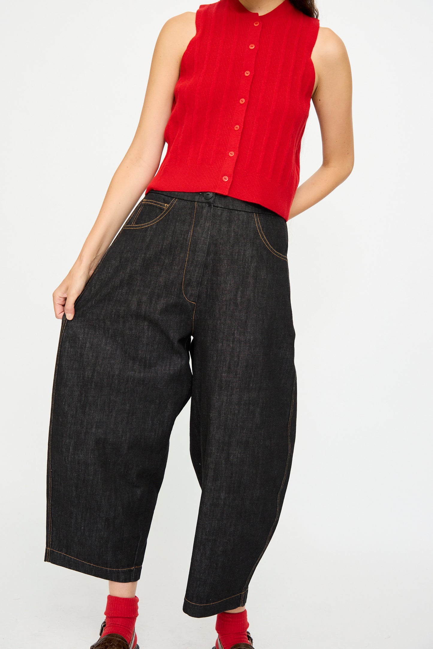 A person wearing a sleeveless red button-up top, Cordera Denim Curved Pant in Black with an elastic waistband, and red socks stands against a plain white background.