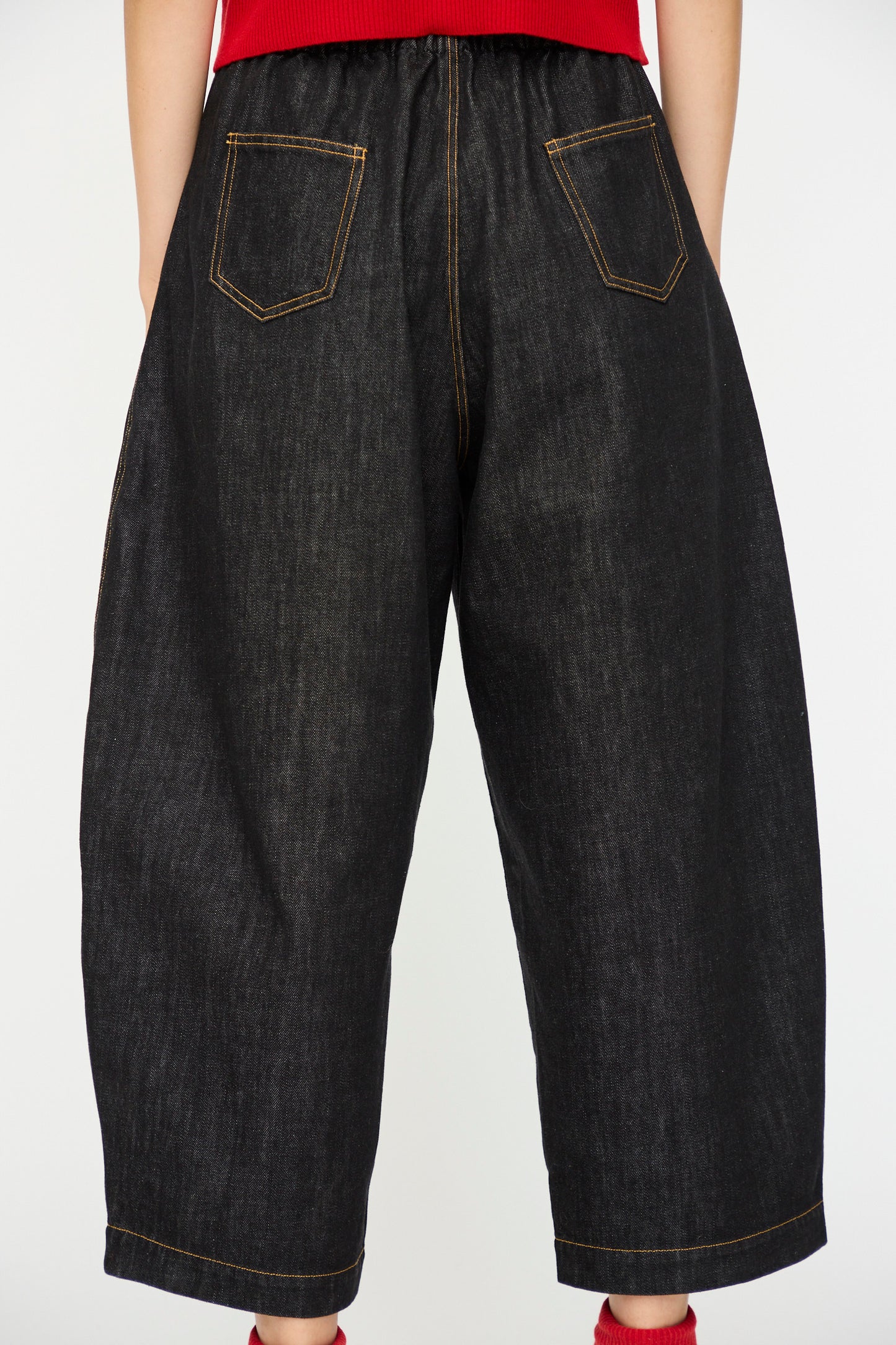 A person wears Cordera's Denim Curved Pant in Black—loose-fitting, dark organic cotton denim jeans with an elastic waistband and yellow stitching—standing against a plain white background.