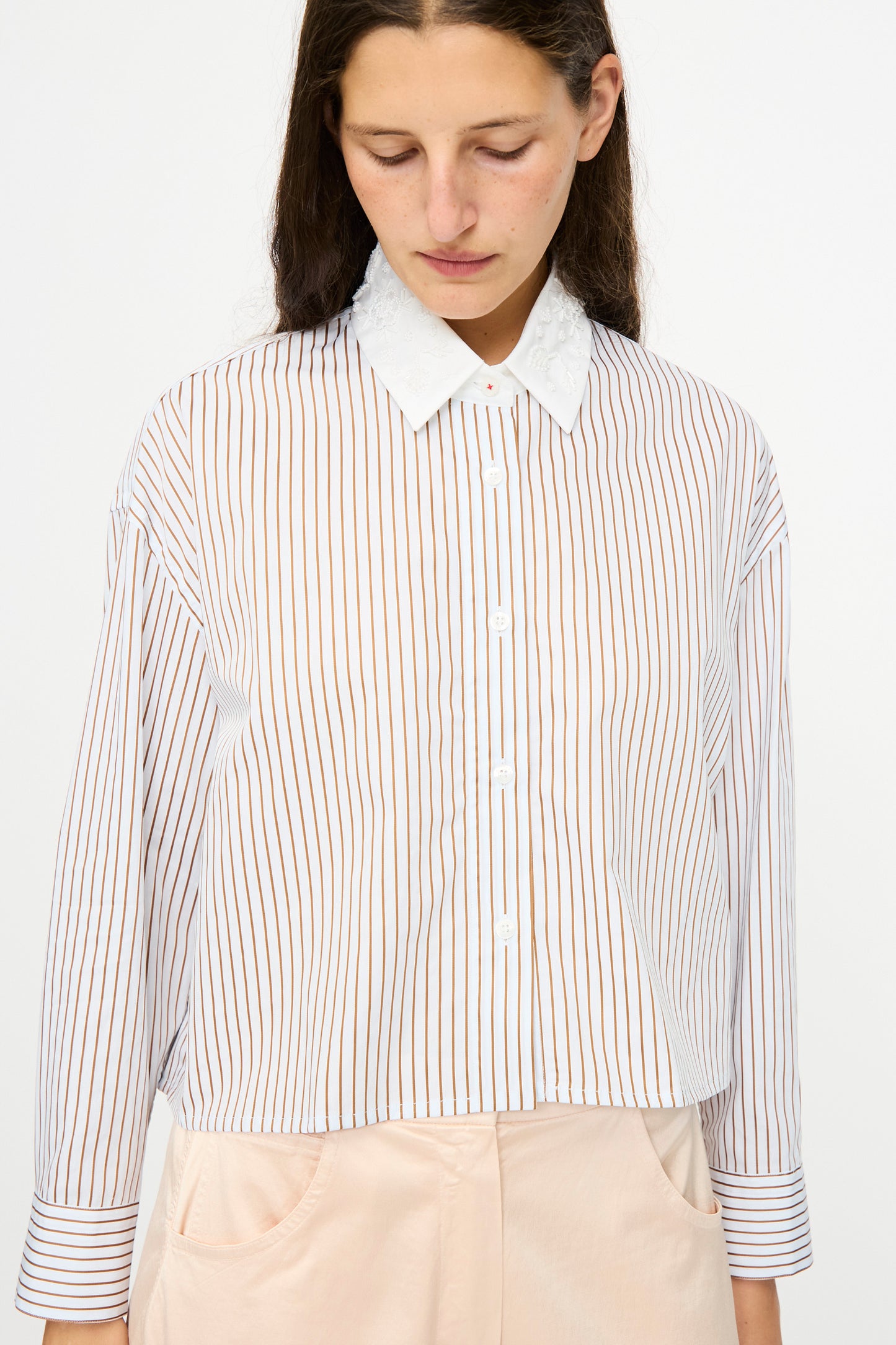 A woman with long brown hair models Cordera's Embellished Shirt in Striped—a white cotton shirt with thin vertical brown stripes—paired with light pants and set against a plain background.