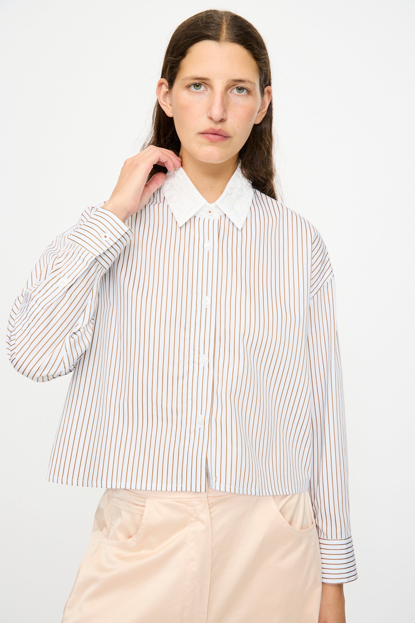 A person with long brown hair wears Cordera's Embellished Shirt in Striped, featuring vertical red and blue stripes, paired with pale pink pants, standing against a plain light background.