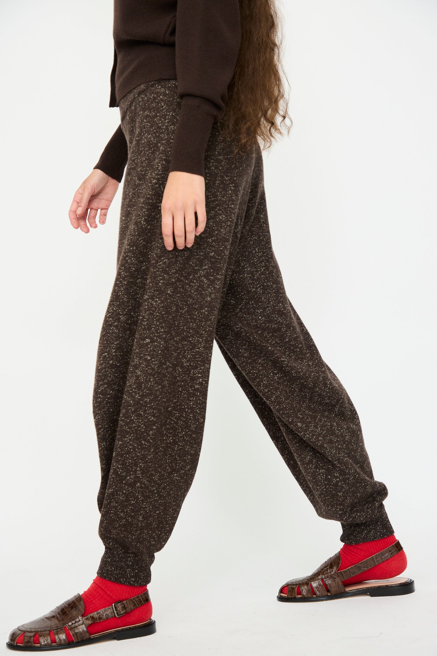 A person wears Cordera’s Flecked Cotton Pant in Brown, paired with a dark brown sweater, red socks, and brown sandals while walking against a plain white background.