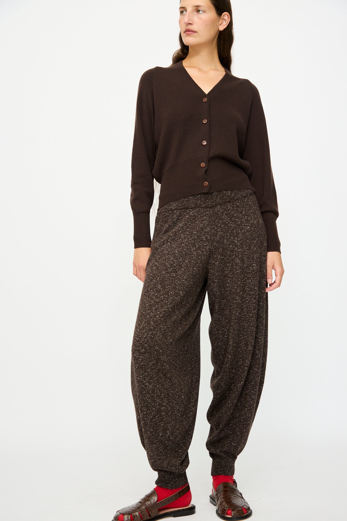 A woman stands against a plain background wearing a dark brown buttoned cardigan, the Cordera Flecked Cotton Pant in Brown, red socks, and brown sandals.