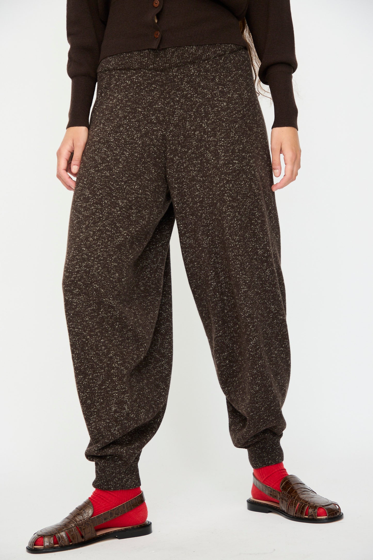 A person wears Cordera's Flecked Cotton Pant in Brown, paired with a dark top, red socks, and brown sandals, standing against a plain white background.