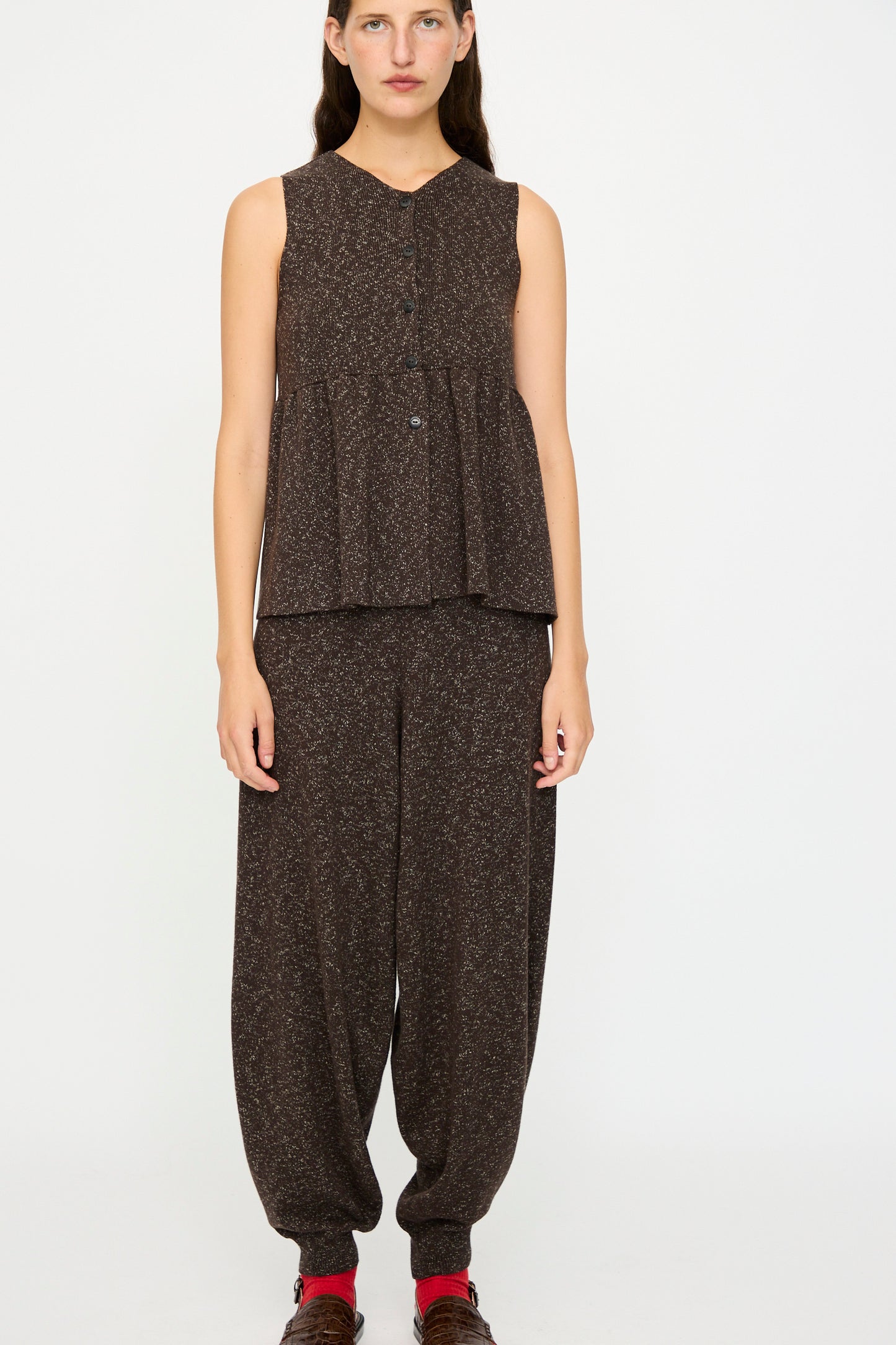 A woman stands against a white background wearing Cordera's Flecked Cotton Peplum Top in Brown, styled sleeveless and buttoned-up, paired with matching loose brown pants, brown shoes, and red socks.