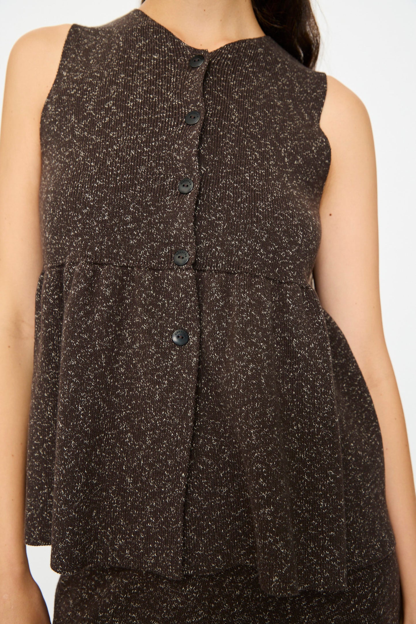 A person wears the Cordera Flecked Cotton Peplum Top in Brown—a dark brown, sleeveless button-up with a speckled pattern and loose, layered peplum silhouette—shown from shoulders to hips.