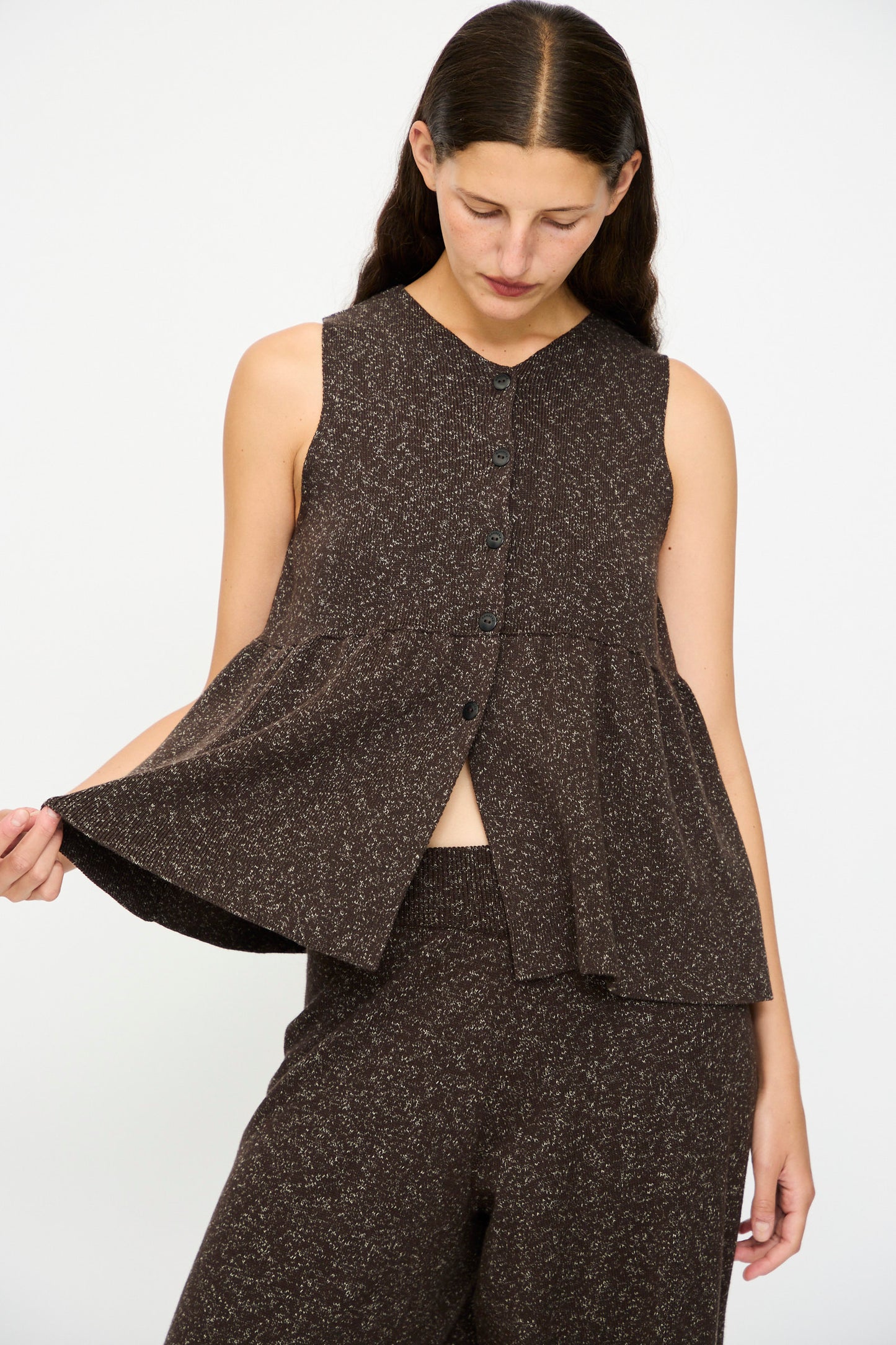 A person wears the Cordera Flecked Cotton Peplum Top in Brown, styled with matching pants, standing against a plain background and holding out the top's side.
